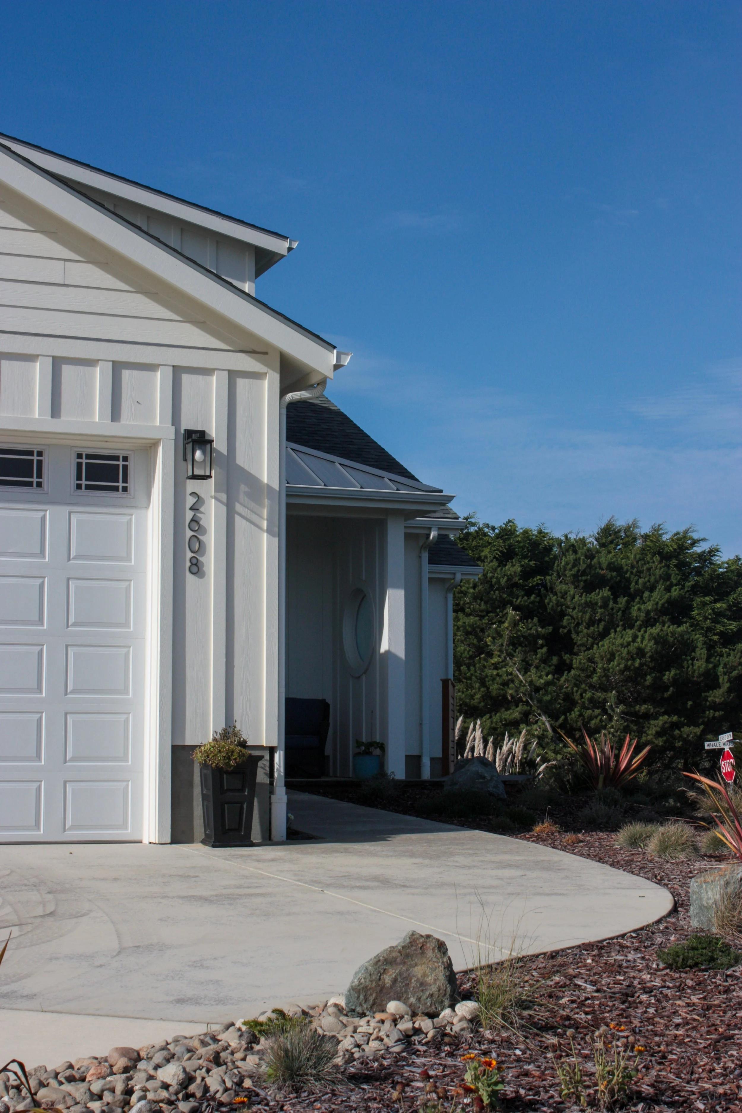 A white modern house with a garage, plant pots, and a curved concrete driveway, set against a blue sky and green trees.