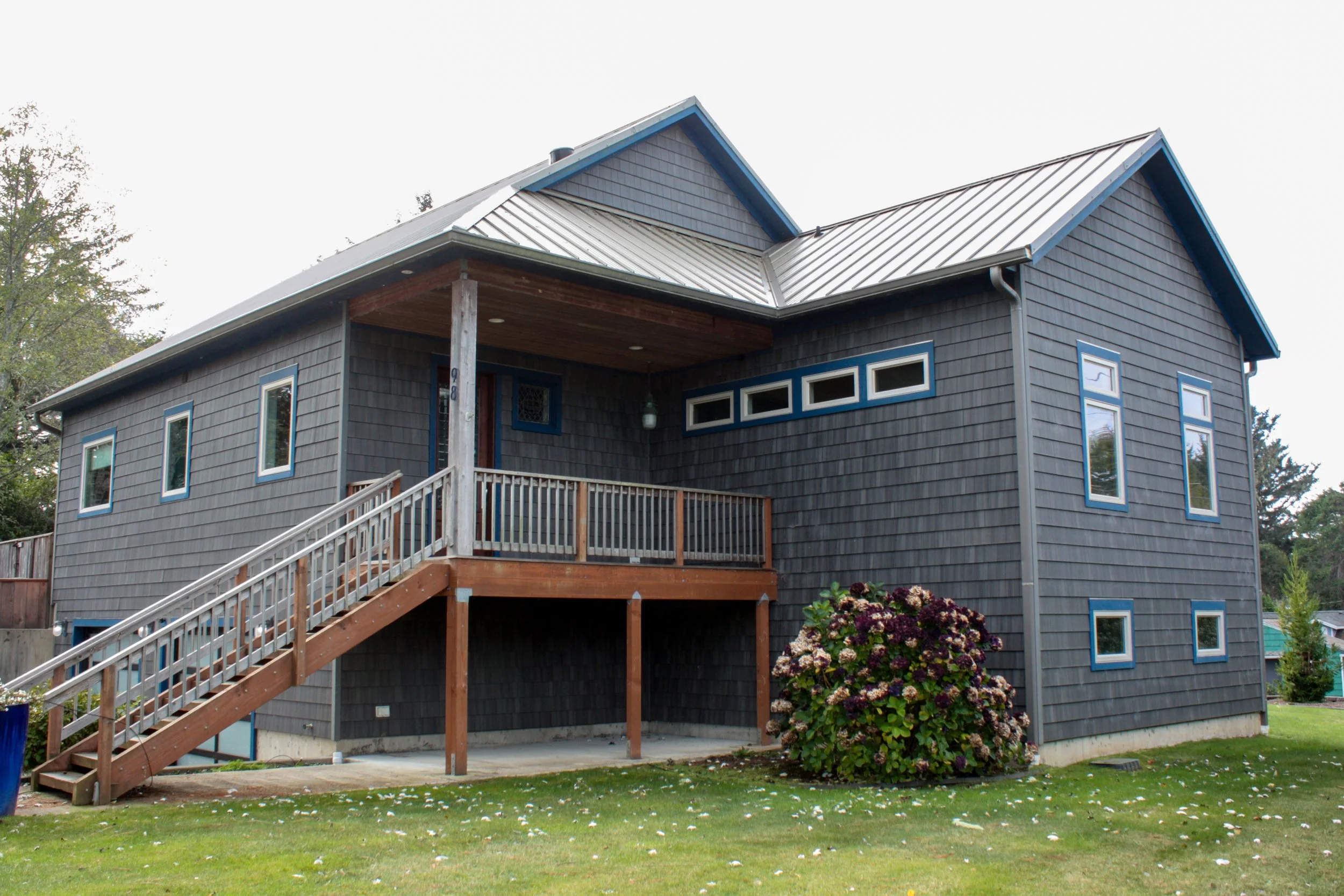 A two-story house with dark gray wooden shingles siding, large windows with blue trim, a metal roof, and a wooden staircase leading to a front porch, with a flowering shrub in the front yard.
