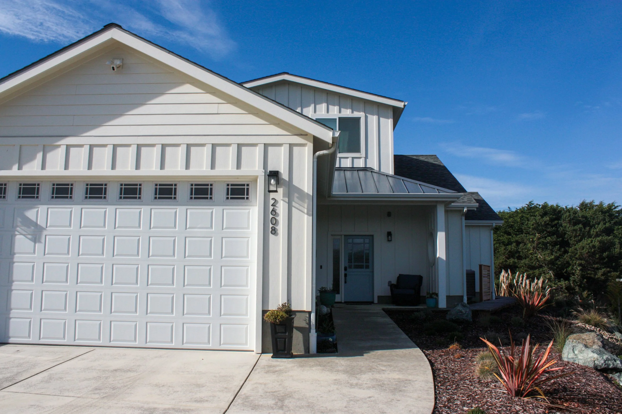 Front view of a modern white house with a garage door, a small porch, and landscaped yard with plants and rocks under a blue sky.