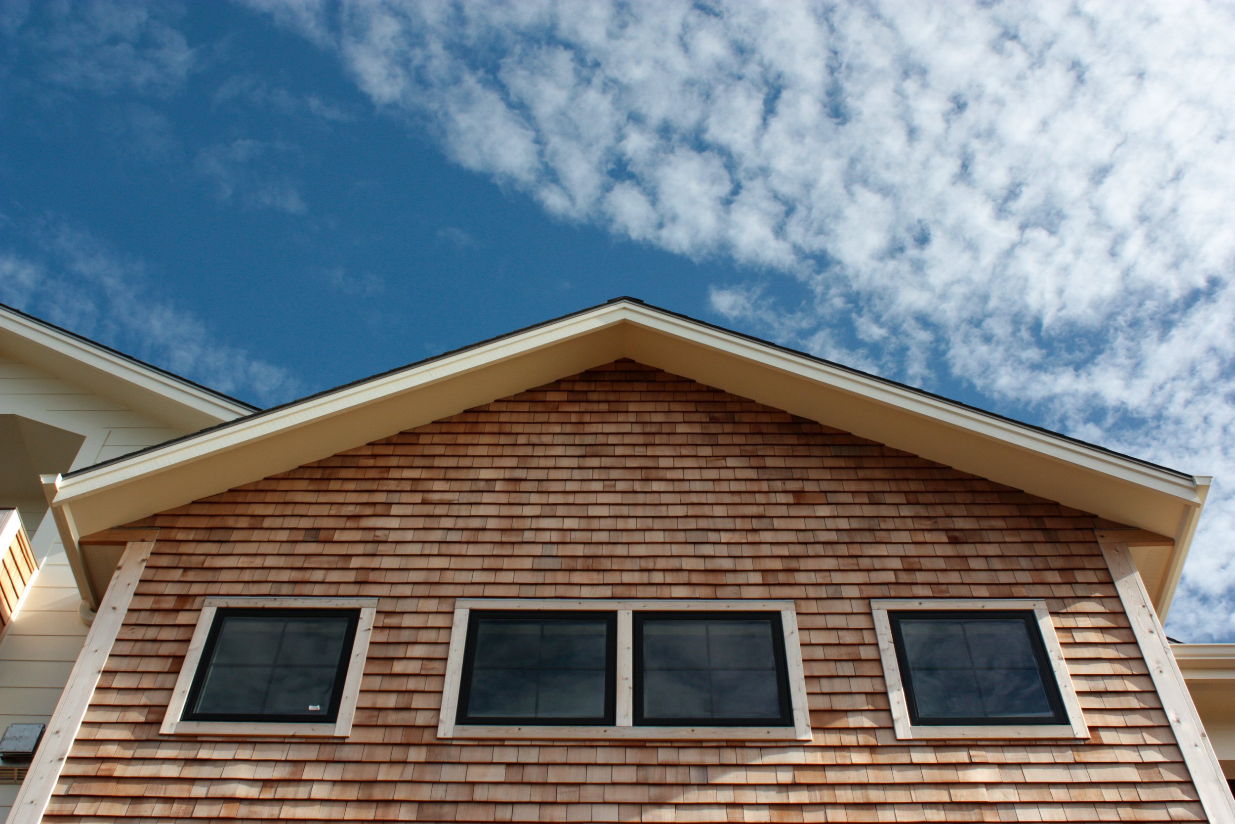 Upward view of a house's gable with brown shingles, three windows, and a blue sky with scattered clouds.