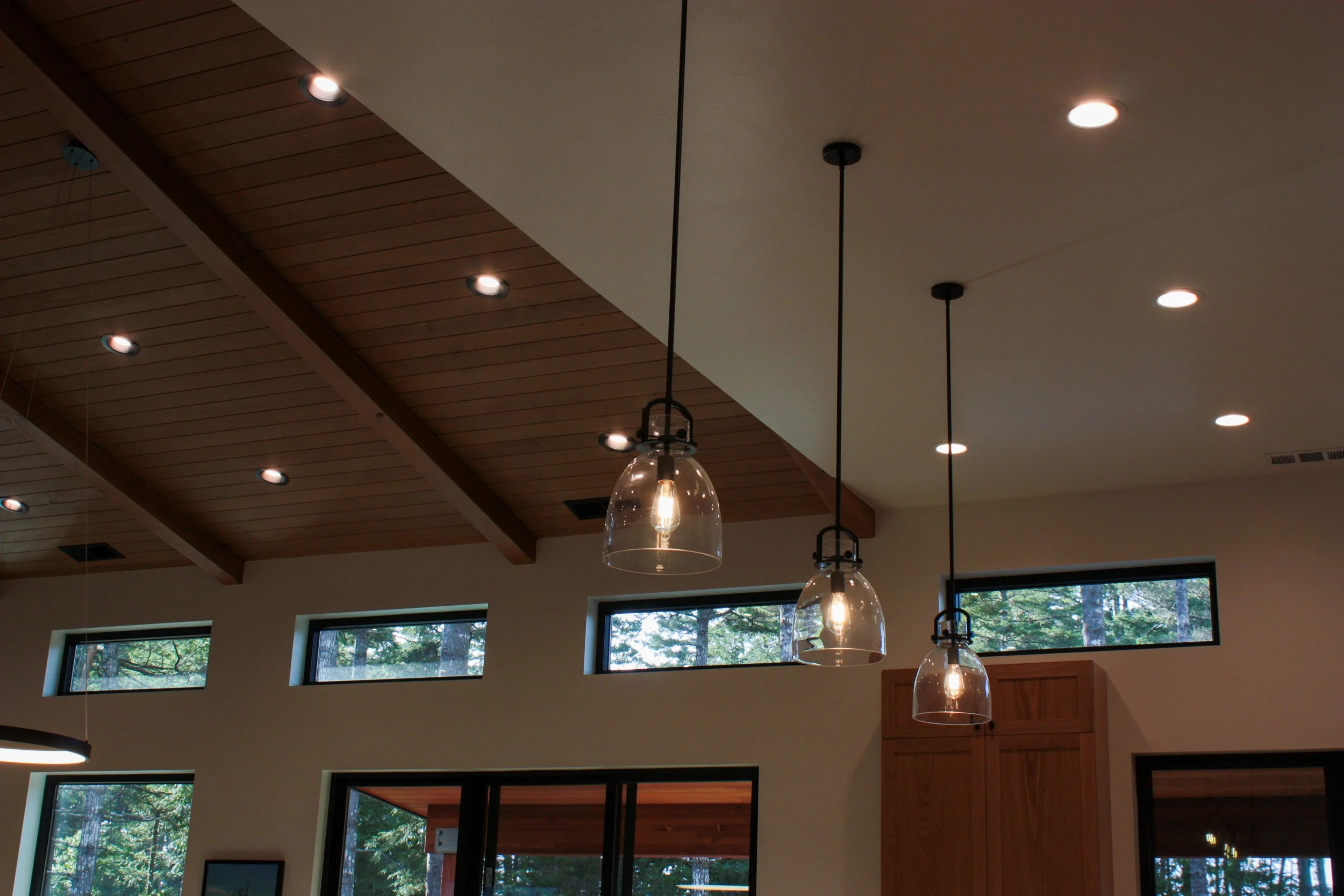 Interior of a modern house with a wooden ceiling, multiple recessed lights, three hanging glass pendant lights with Edison bulbs, and high horizontal windows showing trees outside.