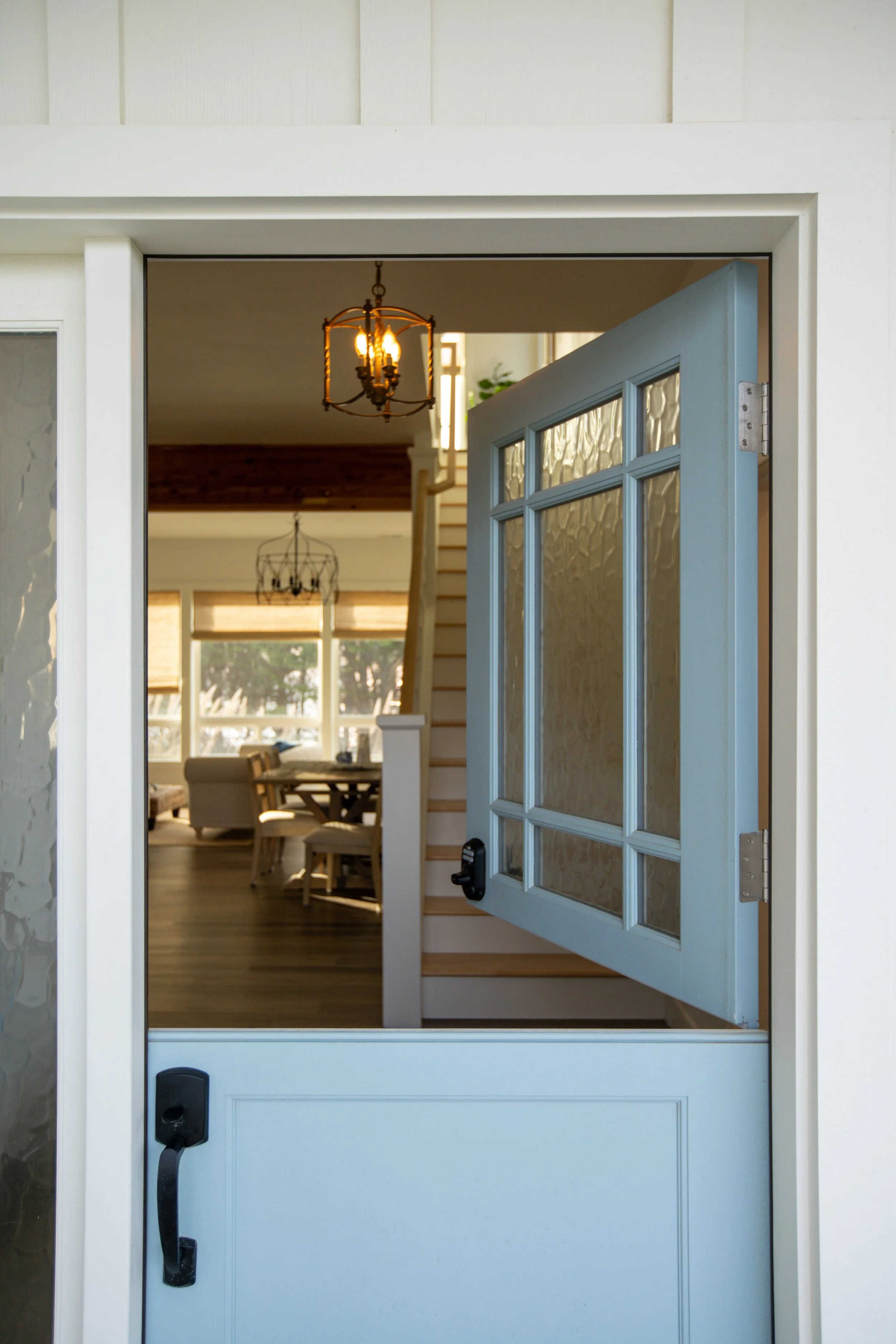 Open front door leading into a dining area with hanging chandeliers, wooden floors, and large windows.