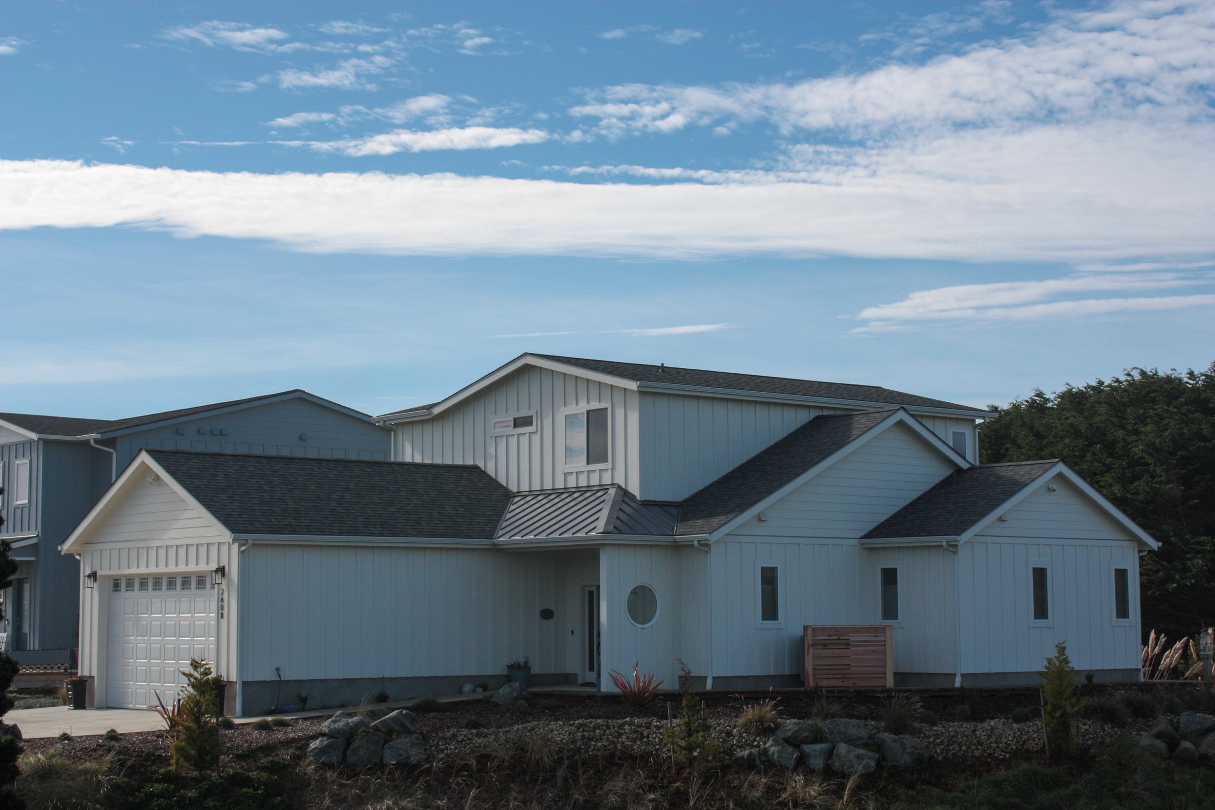 A modern white two-story house with a black roof and attached garage, surrounded by a landscaped yard with rocks and small plants, under a blue sky with scattered clouds.