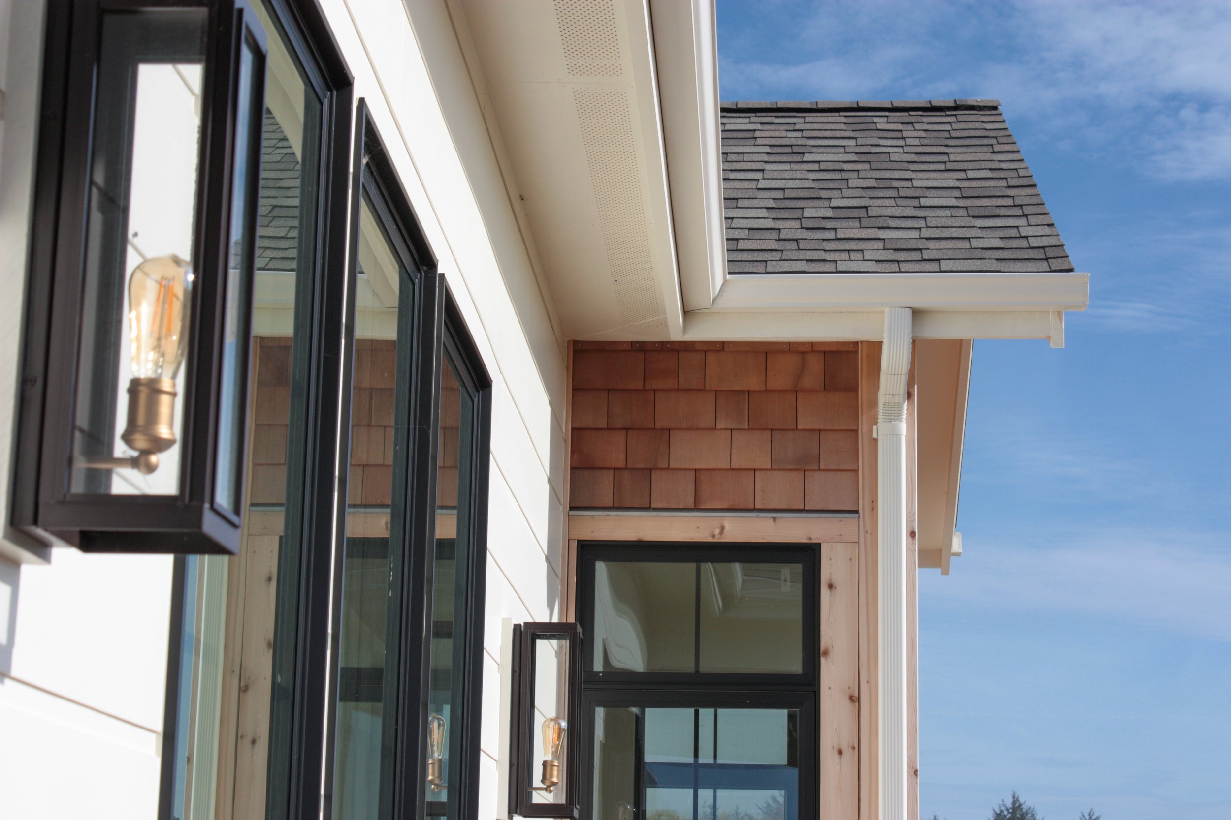 Close-up of a house exterior showing black-framed windows, white siding, and a brown shingled section under a blue sky.
