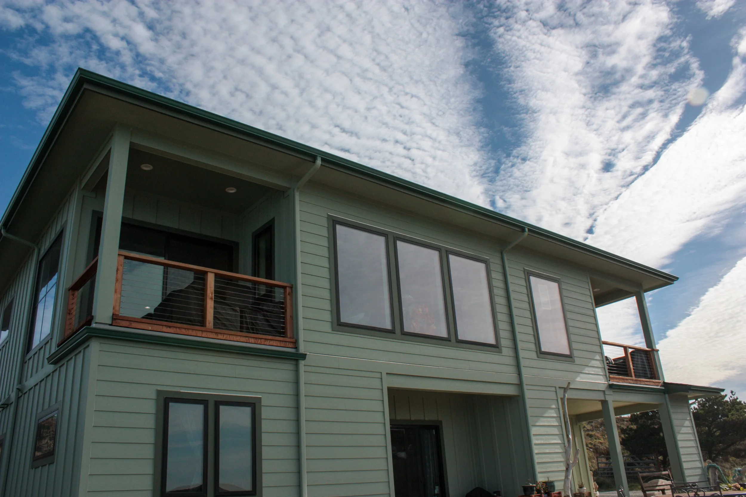 A two-story house with light green siding, black window frames, and a small upper balcony with a wooden railing. The house has a sloped roof and is set against a partly cloudy sky.