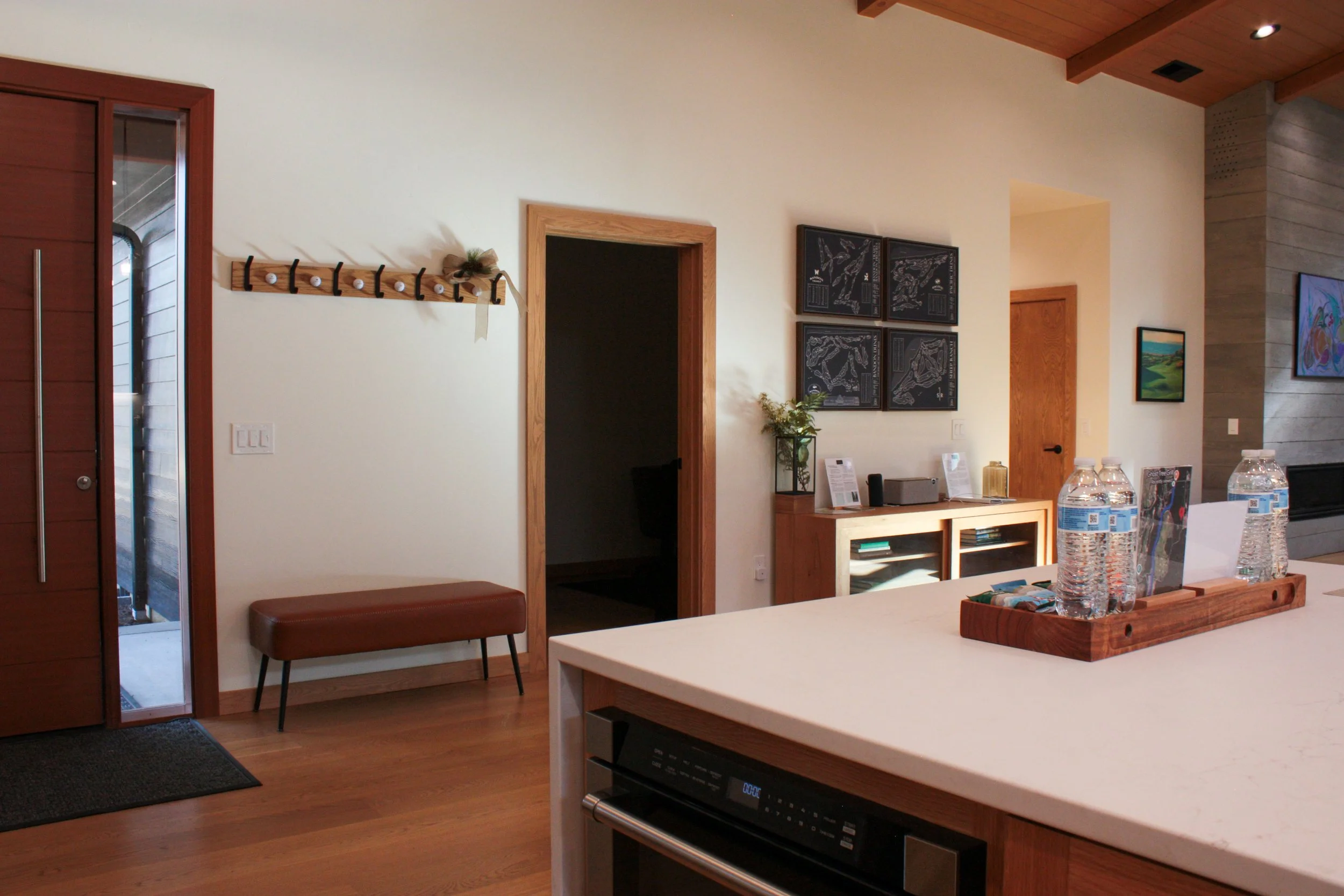 A modern kitchen and living space with wooden accents. There is a white countertop with bottled water, a bench below a coat rack on the wall, a small cabinet with decor items, and framed artwork on the walls. A doorway leads to another room.