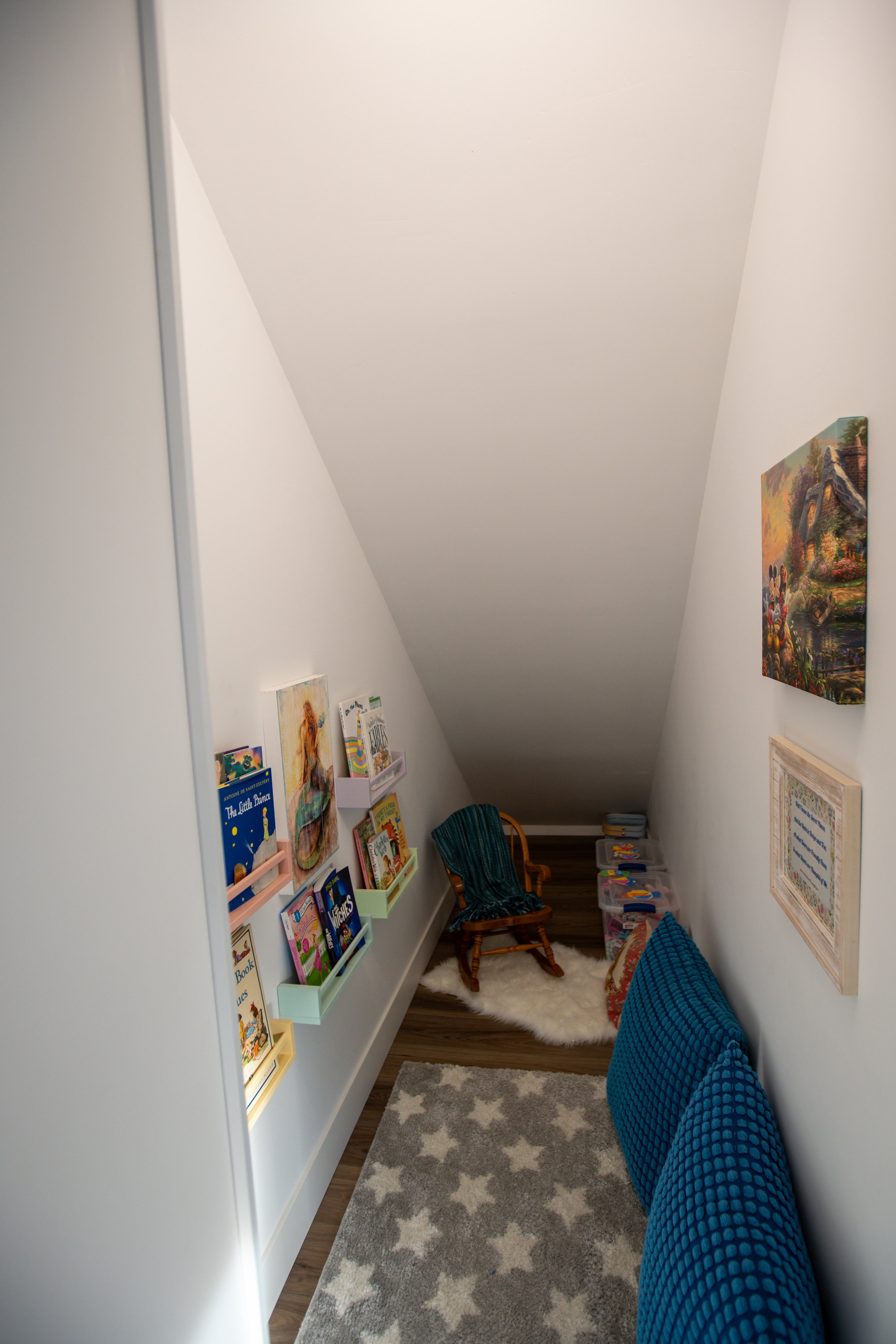 A cozy reading nook with white walls and a sloped ceiling, featuring a wooden rocking chair with a blue blanket, a fluffy white rug, a gray star-patterned mat, and wall-mounted bookshelves filled with children's books.