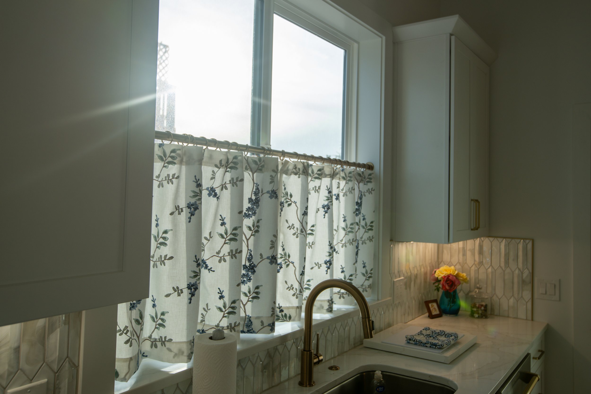 Kitchen window with floral curtains, white cabinets, marble countertop, gold faucet, and a vase with flowers.