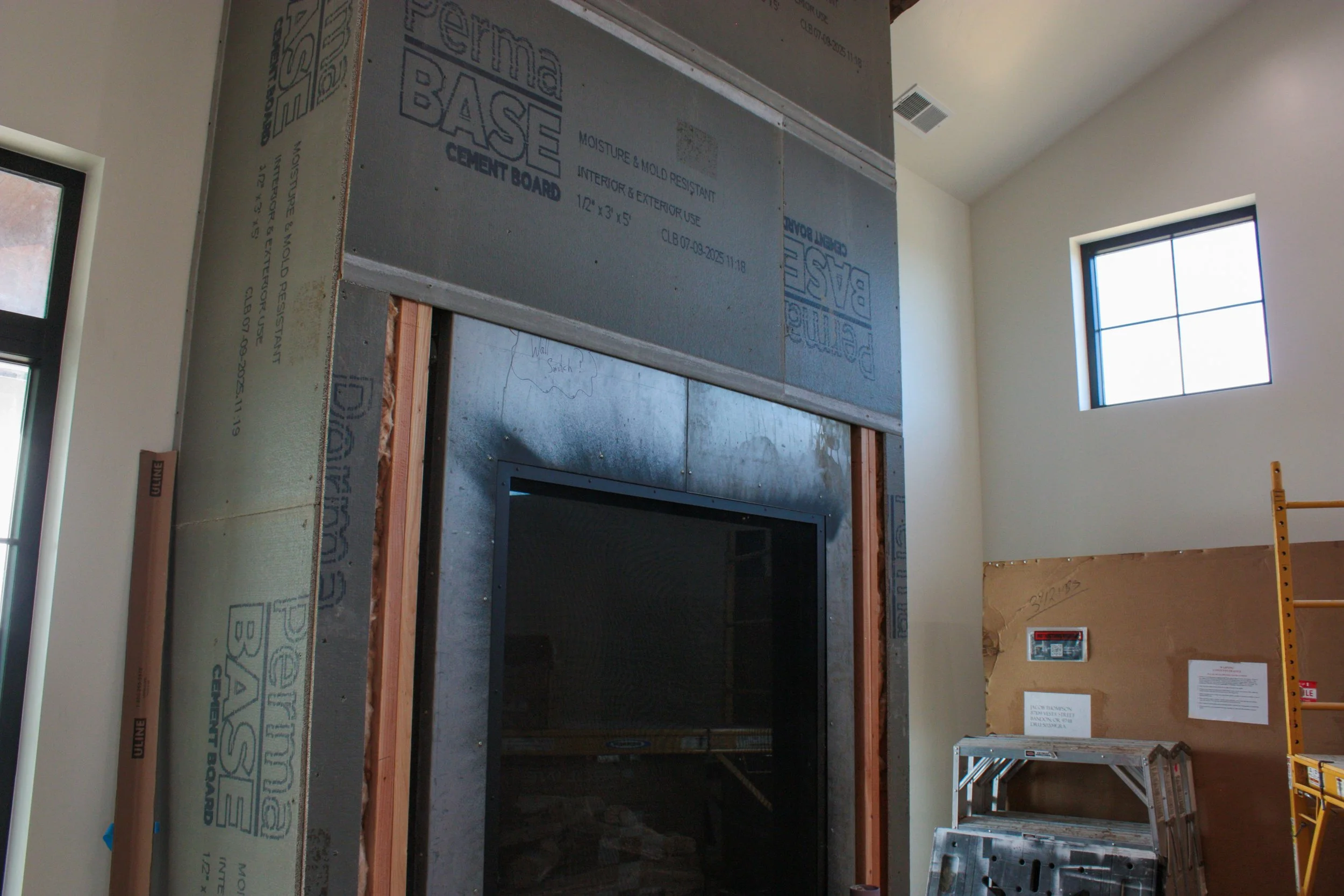 Interior of a building under construction with a fireplace opening, dry wall, scaffolding, and a window.