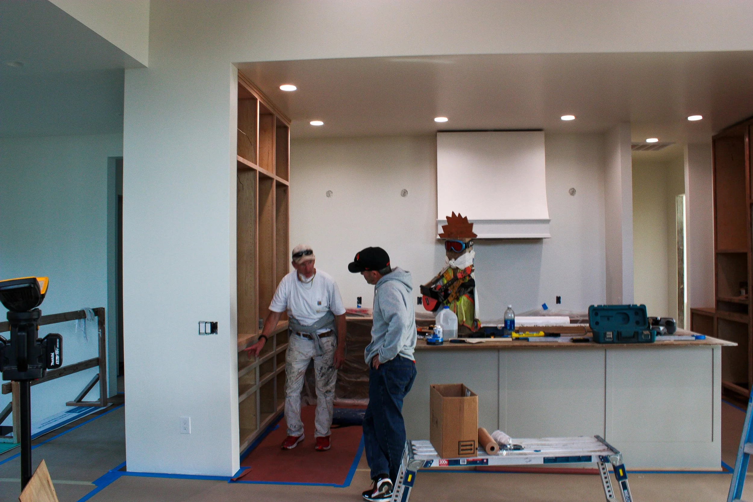 Two construction workers discussing in a partly built kitchen, with cabinets and tools, while a colorful animated figure wearing sunglasses and a wig stands in the background.