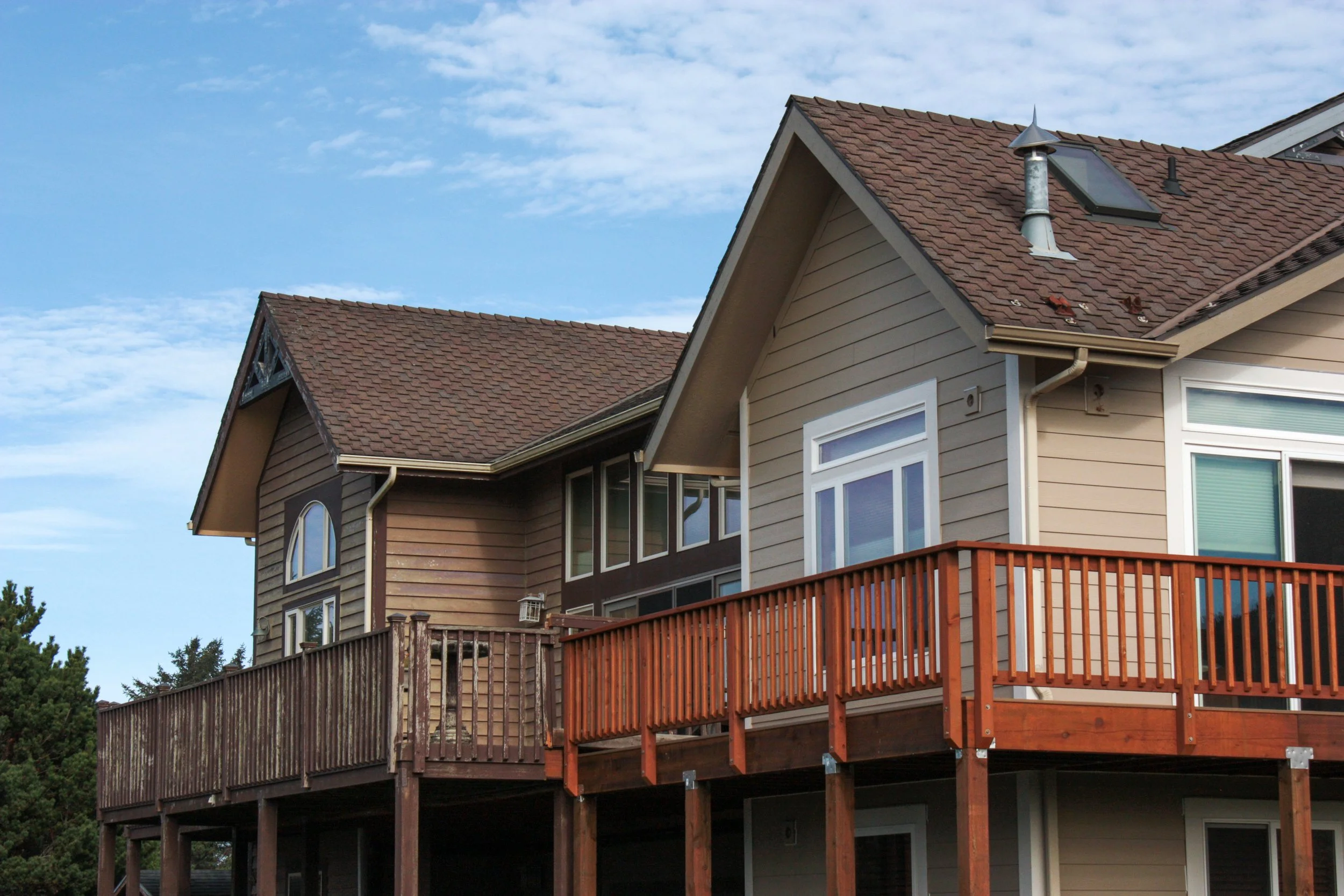 A two-story house with beige and brown siding, a brown shingle roof, and a wooden balcony on the second floor, under a partly cloudy sky.