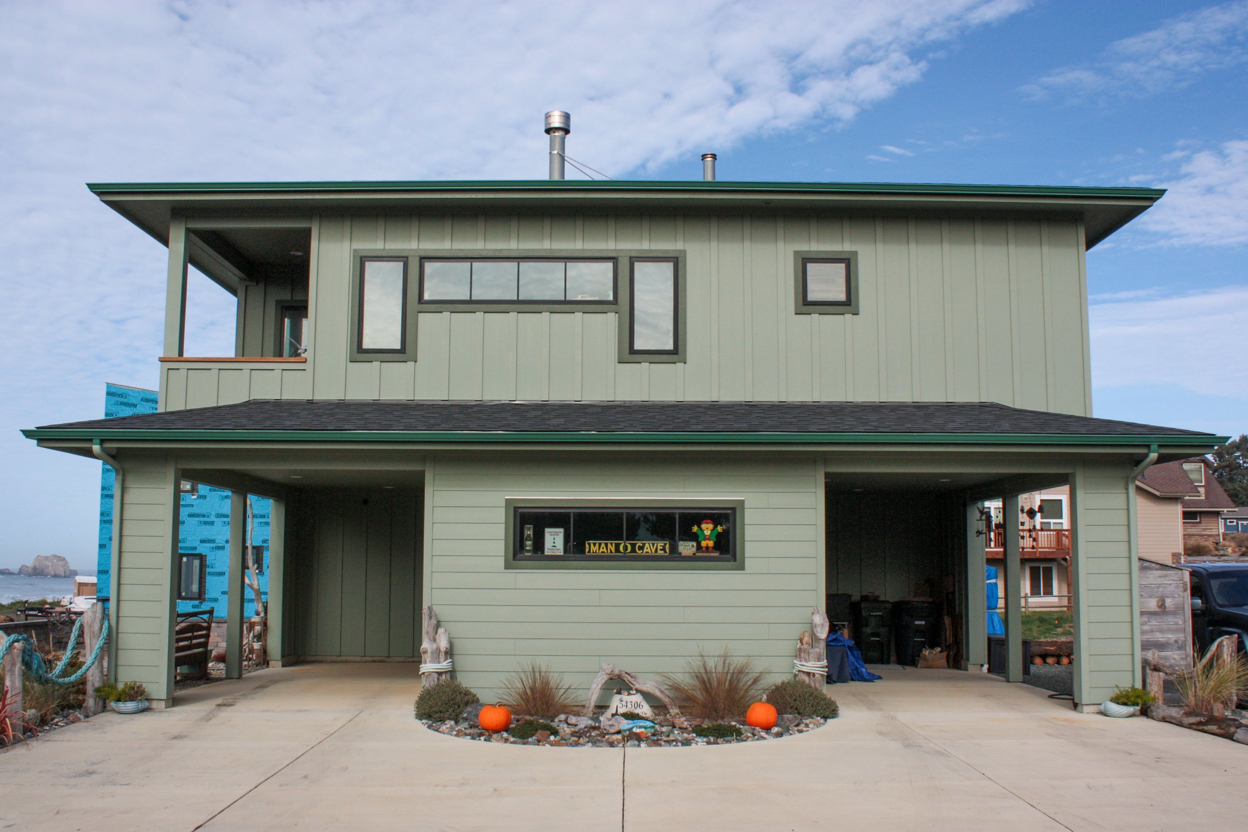 A two-story green house with a balcony on the second floor, a large window, and a small window on the upper level. The garage area on the ground floor is open, with Halloween pumpkins and decorations outside. The driveway is concrete with plants and 