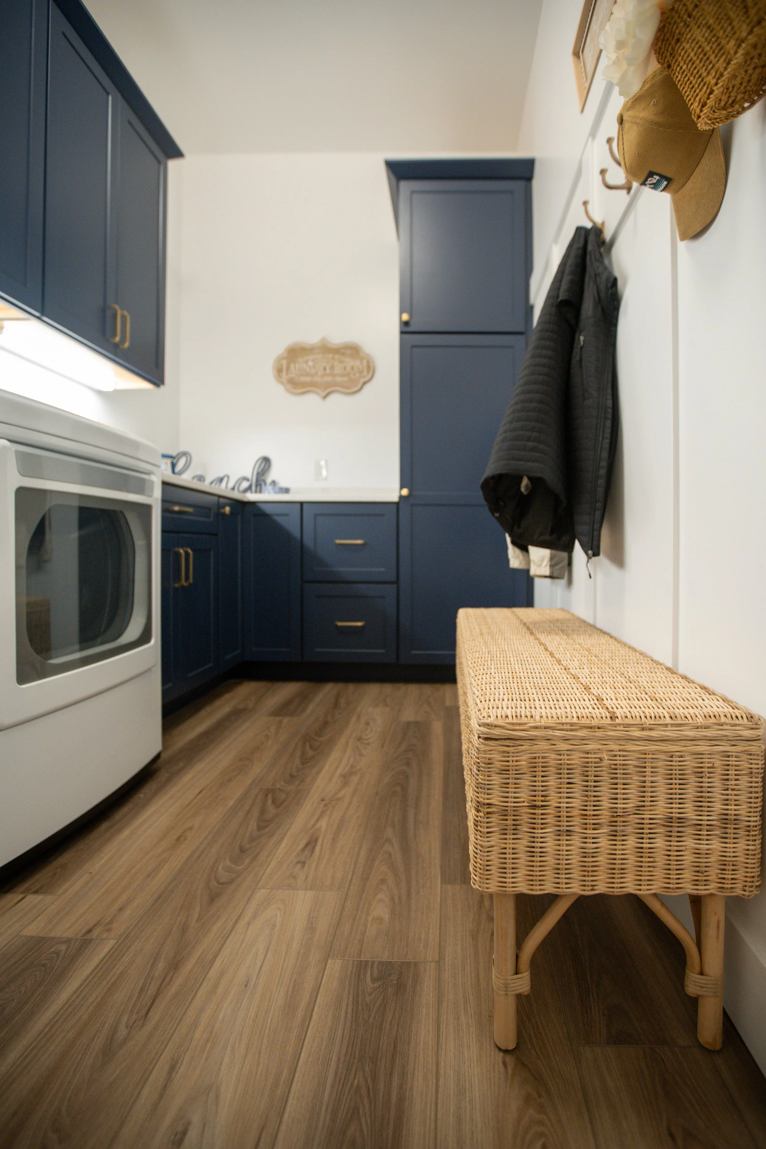 Cozy kitchen with blue cabinets, white walls, wood flooring, a woven bench, hanging jackets, and hats on the wall.