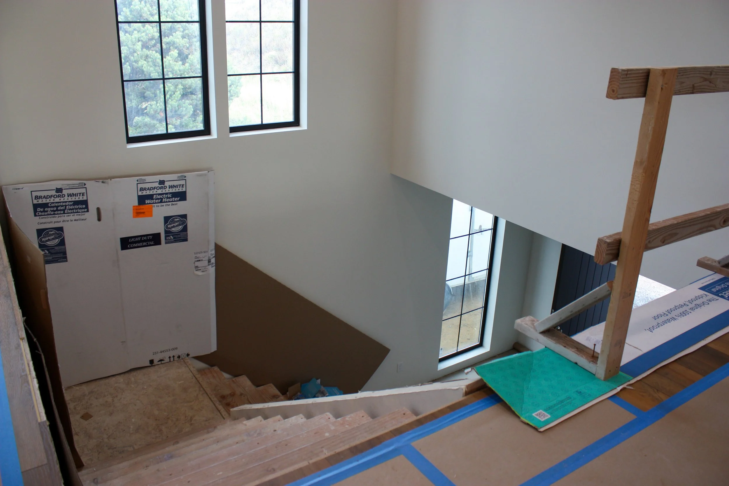 Interior view of a house under construction, showing stairs, large windows, and a boxed water heater in an unfinished hallway.