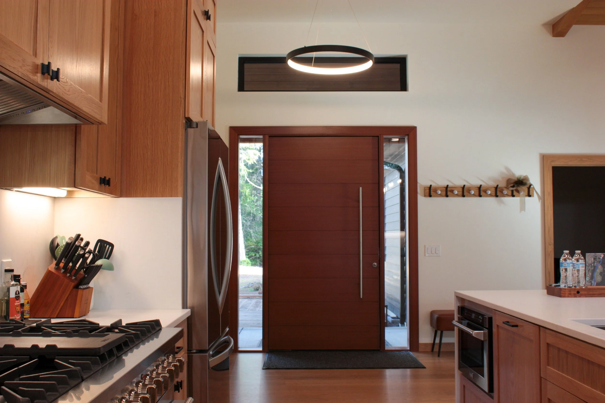 Modern kitchen with wooden cabinets, stainless steel refrigerator, white countertops, and a large wooden front door with glass panels.