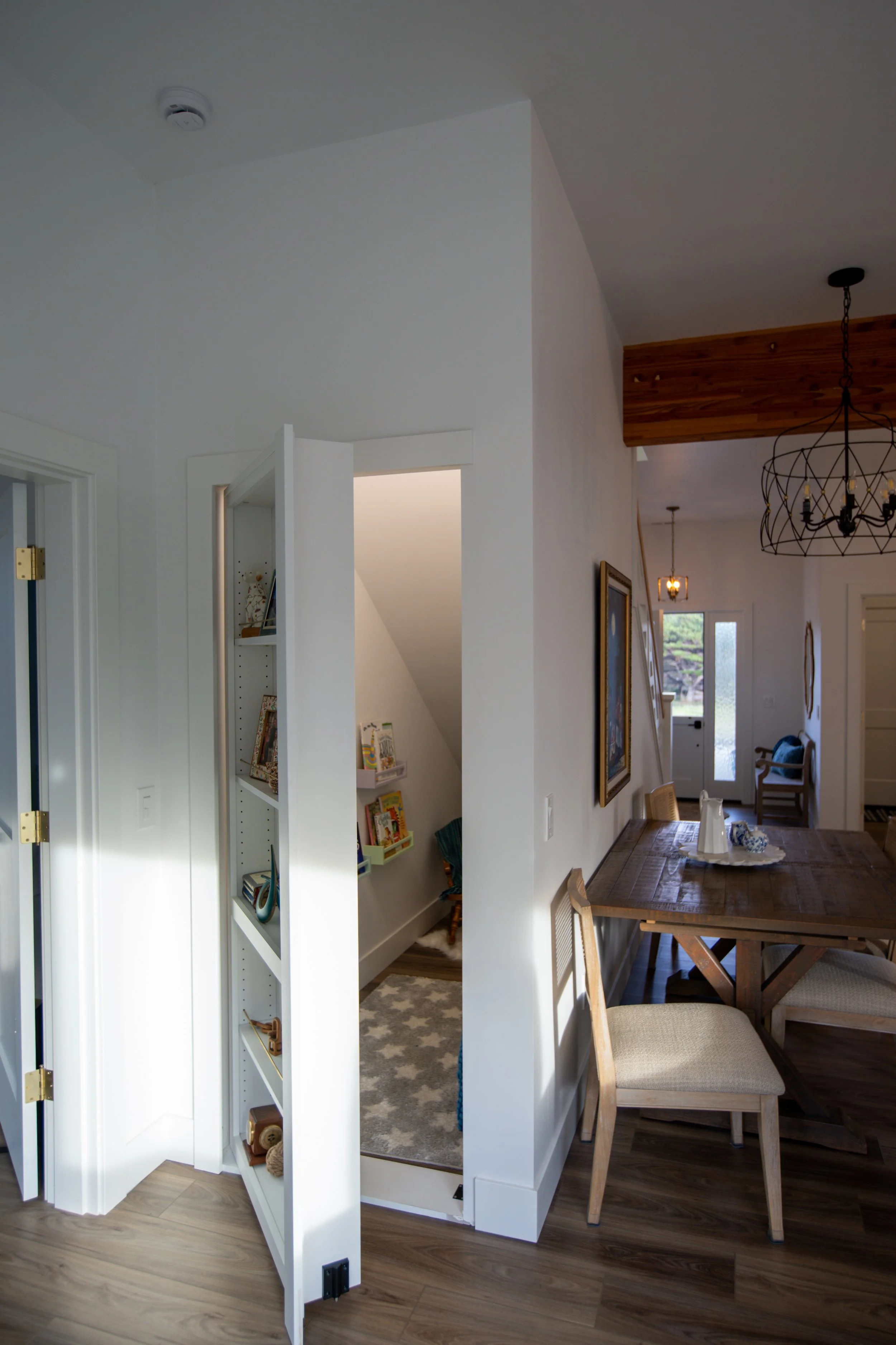 Interior view of a home with a dining table, chairs, artwork on the wall, and a doorway leading to a small room with shelves and picture books.