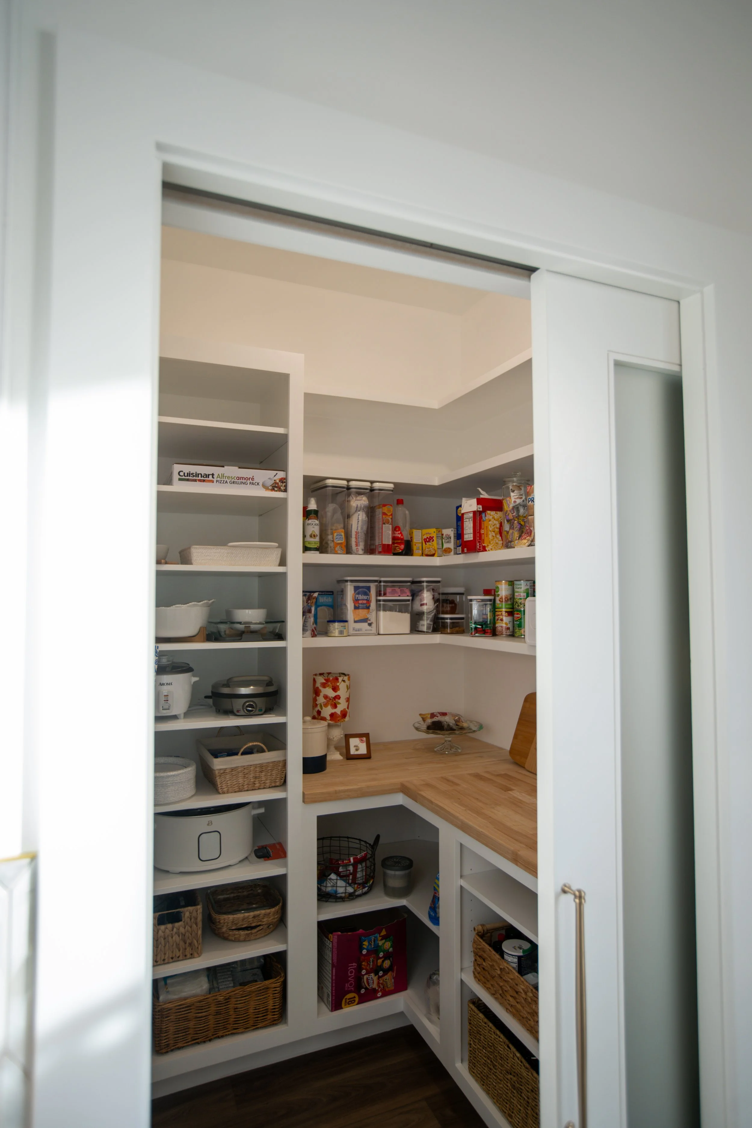 Pantry with white shelves filled with food containers, canned goods, snacks, and kitchen appliances, and a countertop with a floral lamp and framed photo.