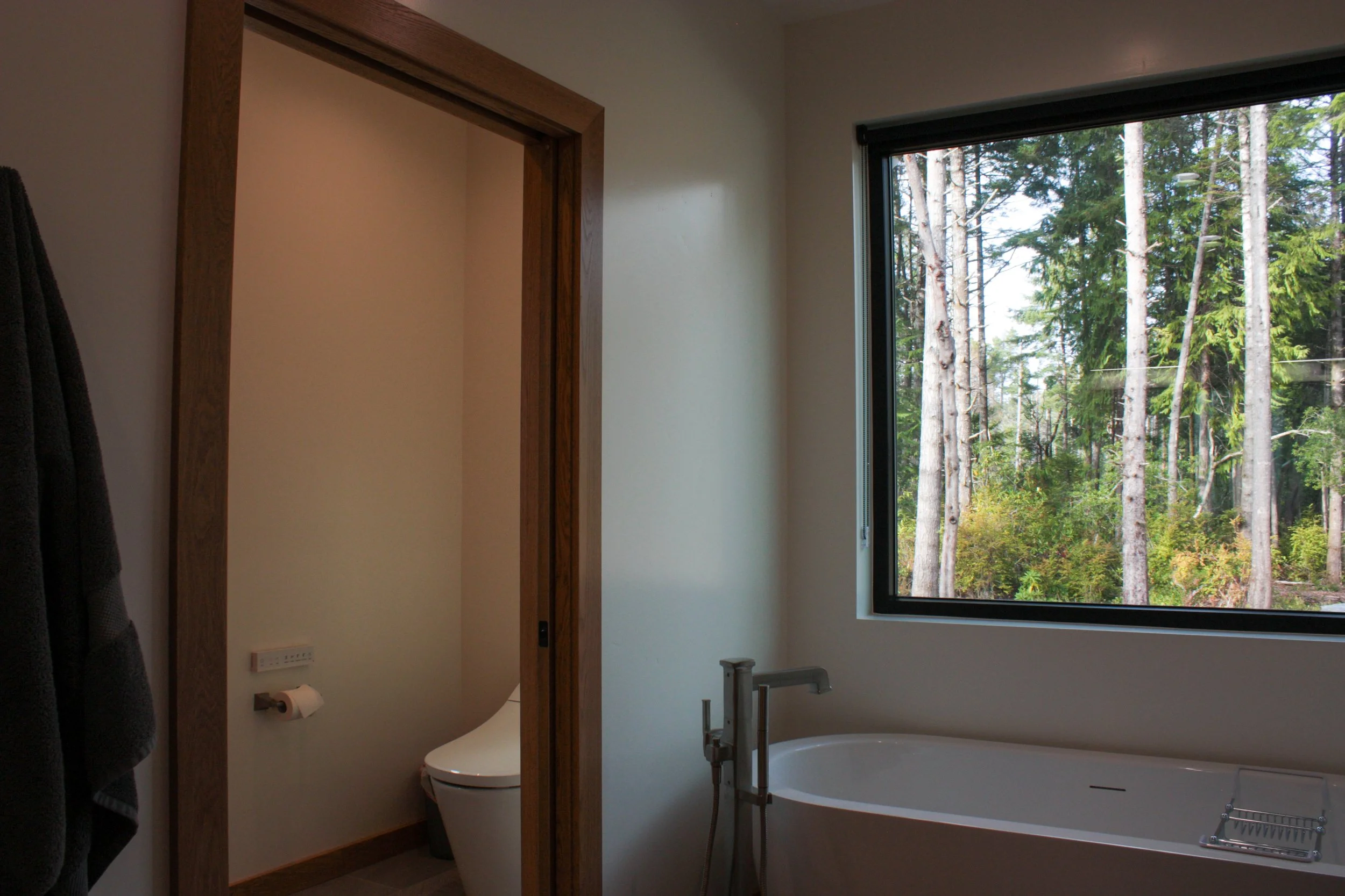 Bathroom with a view of a forest through a large window, a modern bathtub, and a separate toilet room with a toilet.