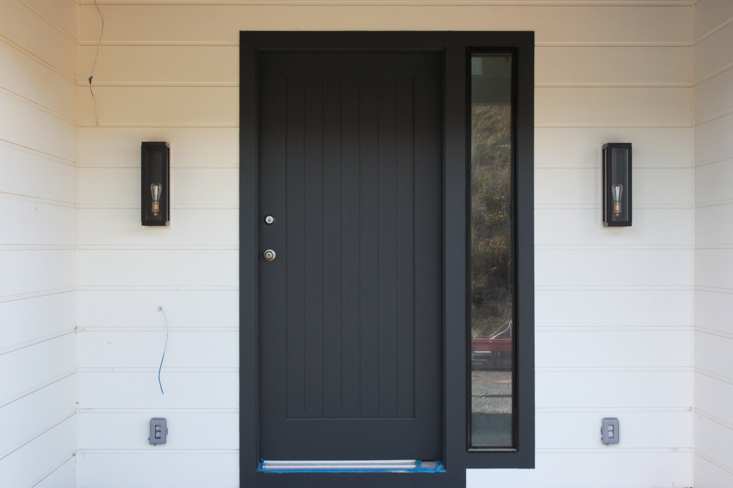 Black front door with a vertical window panel on the right, two black wall-mounted light fixtures on each side, and electrical outlets near the bottom.