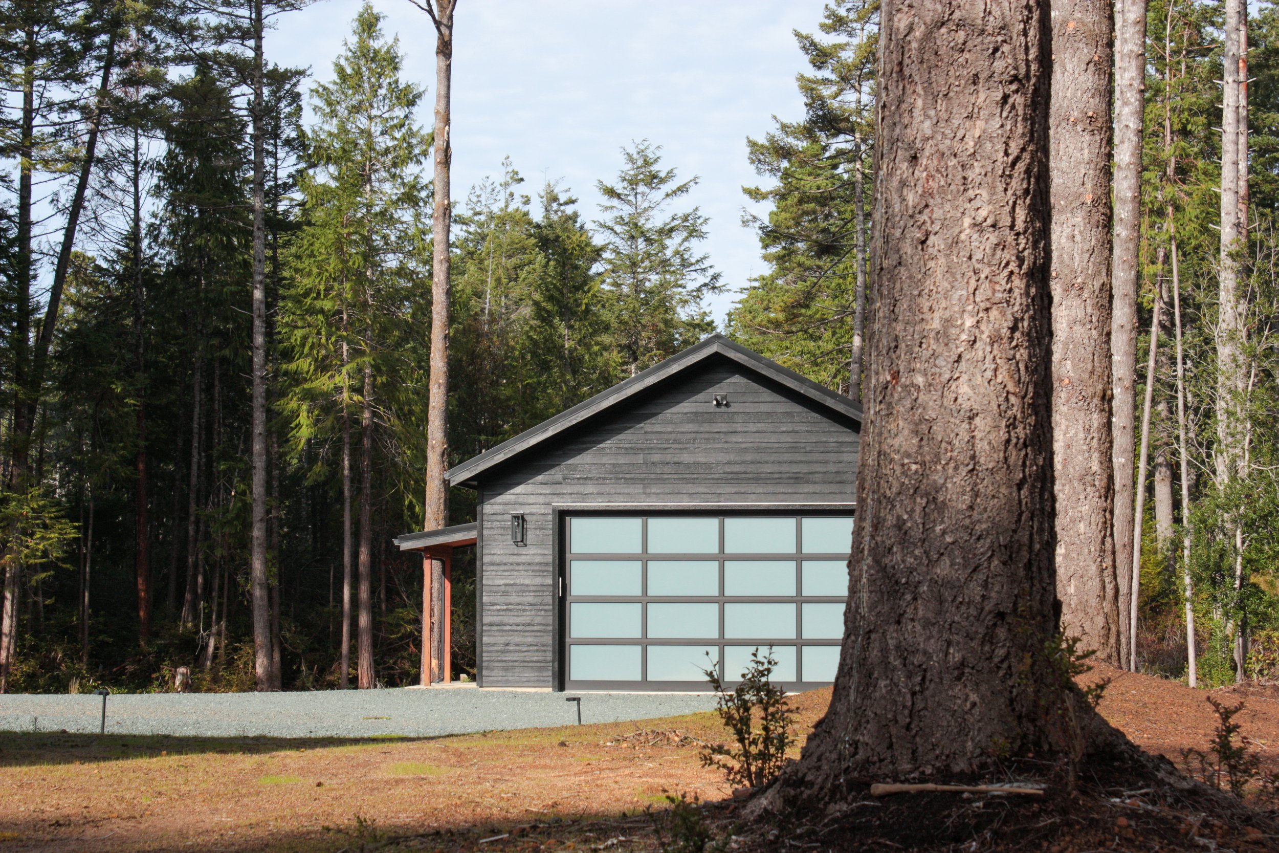 A small, dark gray garage with glass-paneled door, surrounded by tall trees in a forested area, with a gravel driveway in front.