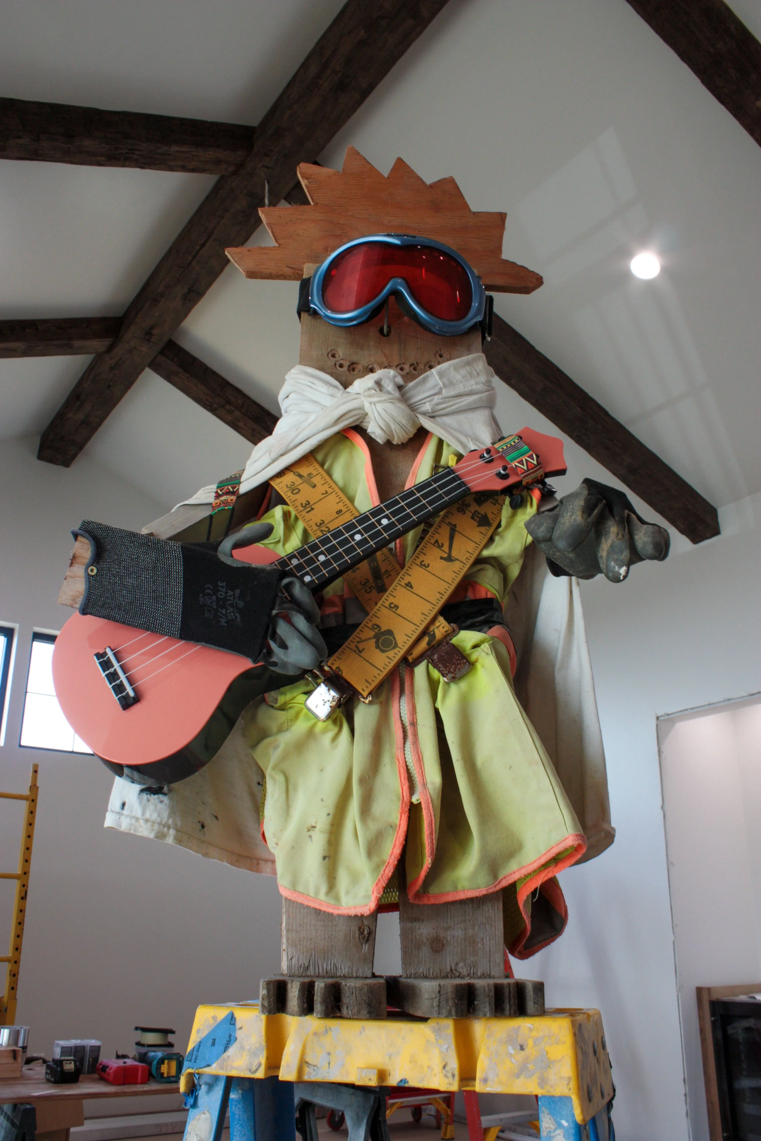 A scarecrow dressed as a construction worker holding a guitar, wearing safety gloves, a measuring tape, and goggles, with a wooden hat and a yellow vest, standing inside a building with exposed wooden beams.