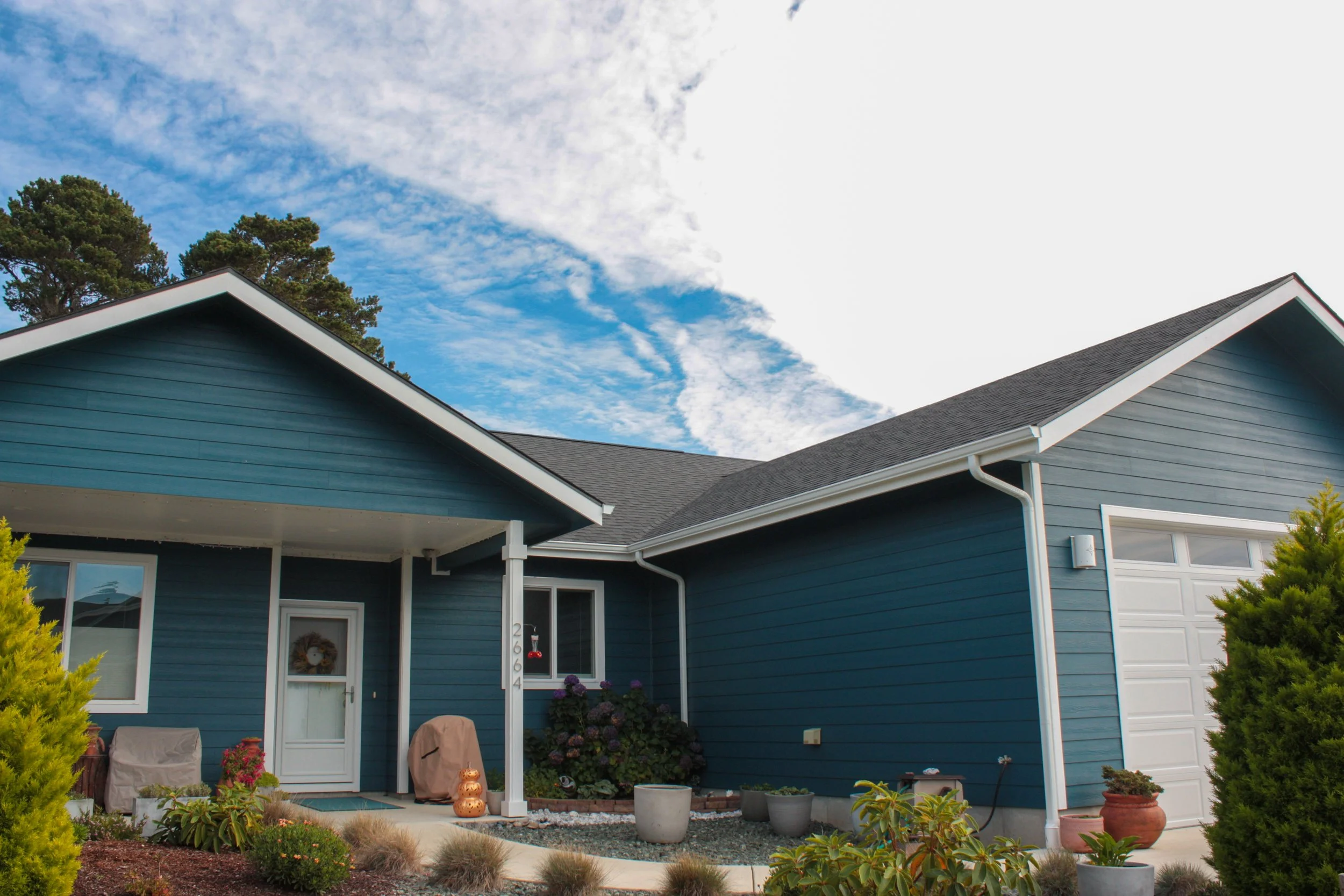 Front view of a blue house with white trim and a garage, surrounded by potted plants and bushes under a partly cloudy sky.