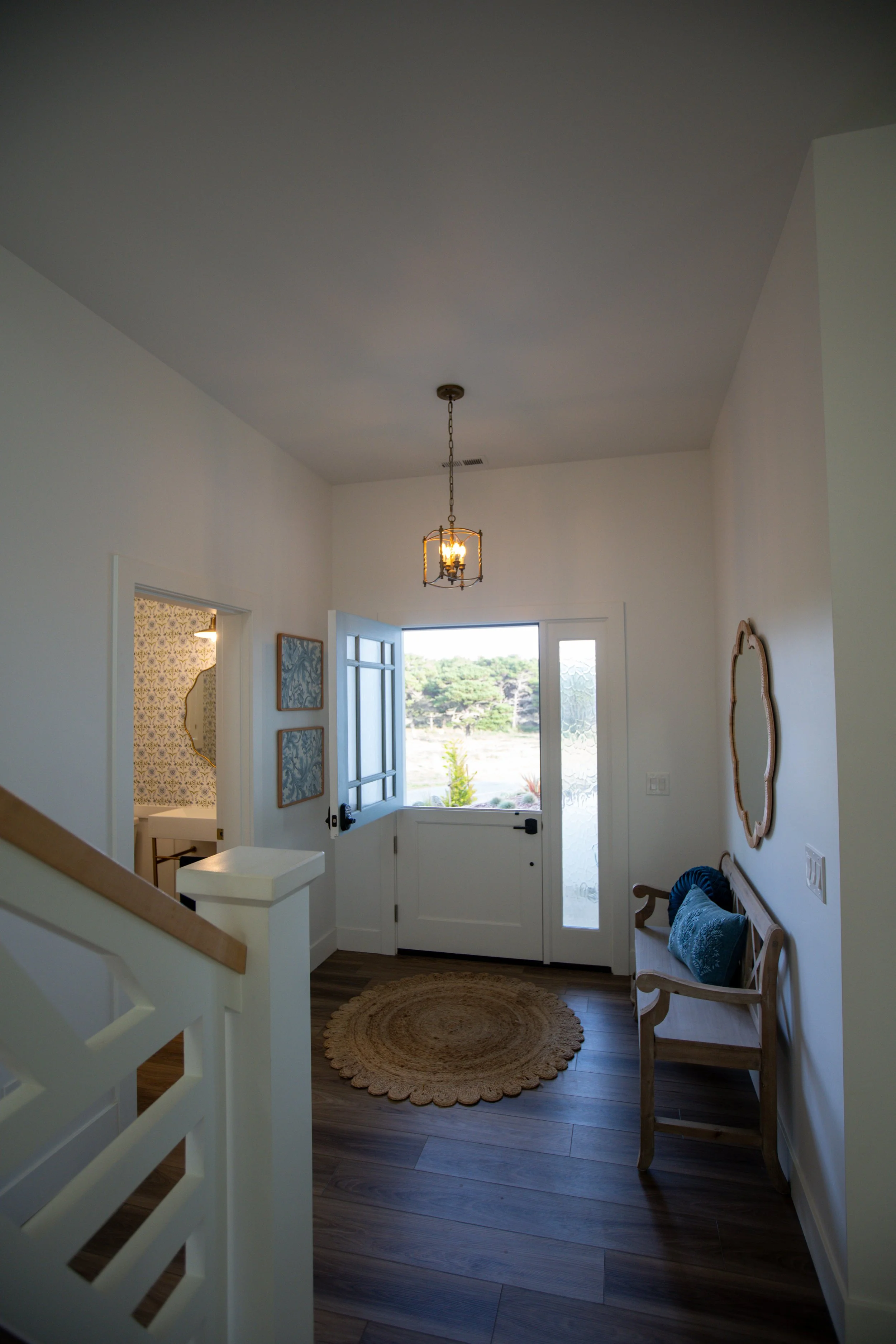 Entryway with open door showing a scenic landscape outside, a bench with cushions, a round woven rug, and wall art in a cozy home interior.