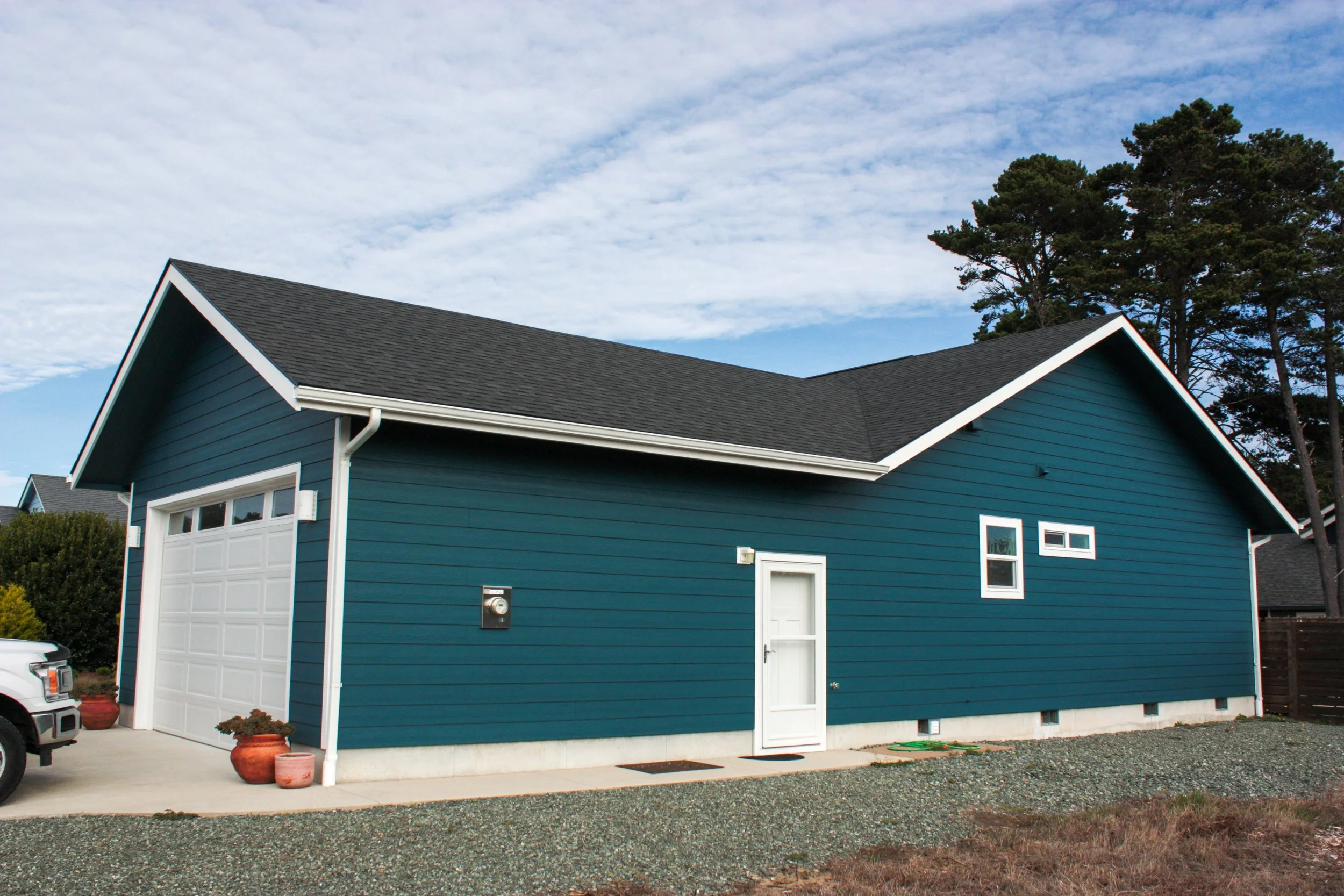 A blue house with white trim and a two-car garage, situated in a suburban setting with trees and a cloudy sky.