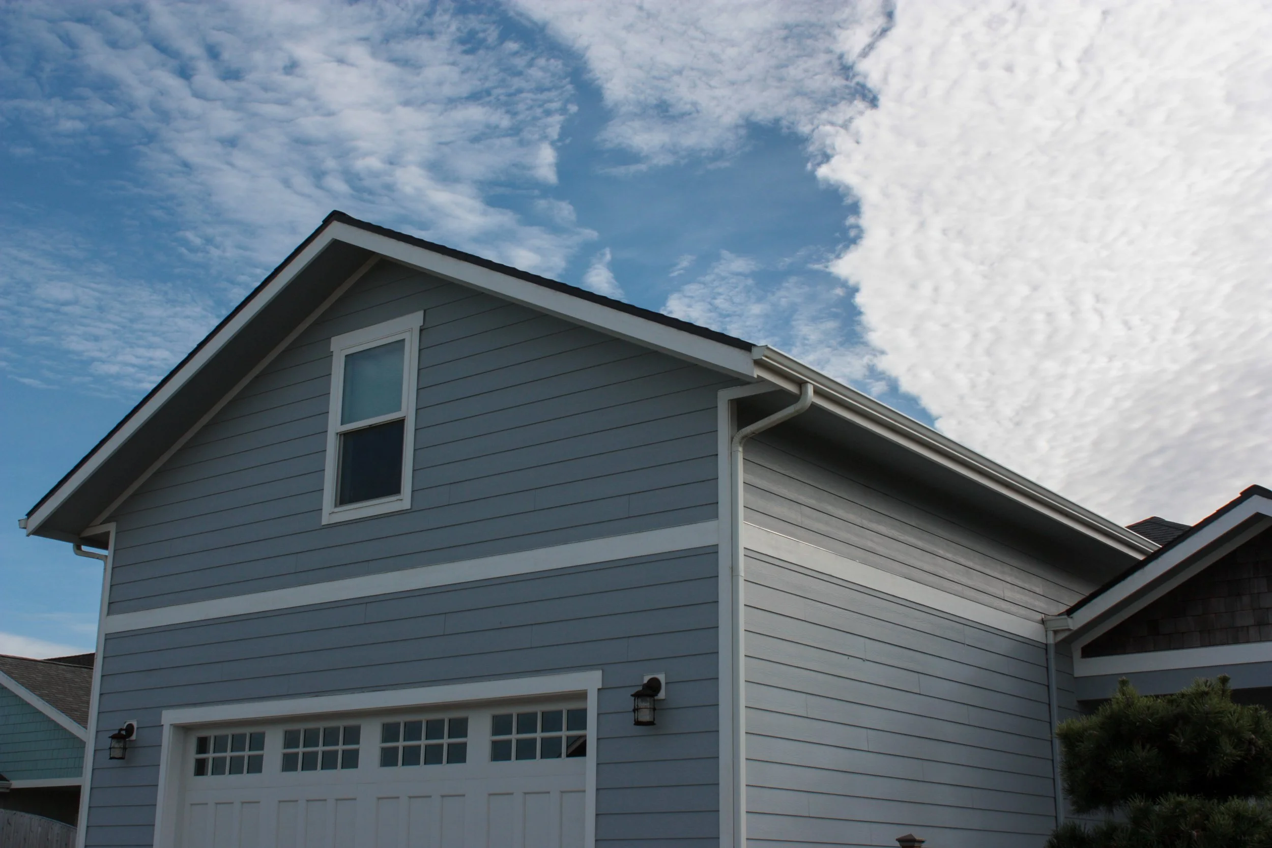 A blue house with a gabled roof and white trim, part of a garage is visible, with a clear sky with some clouds in the background.