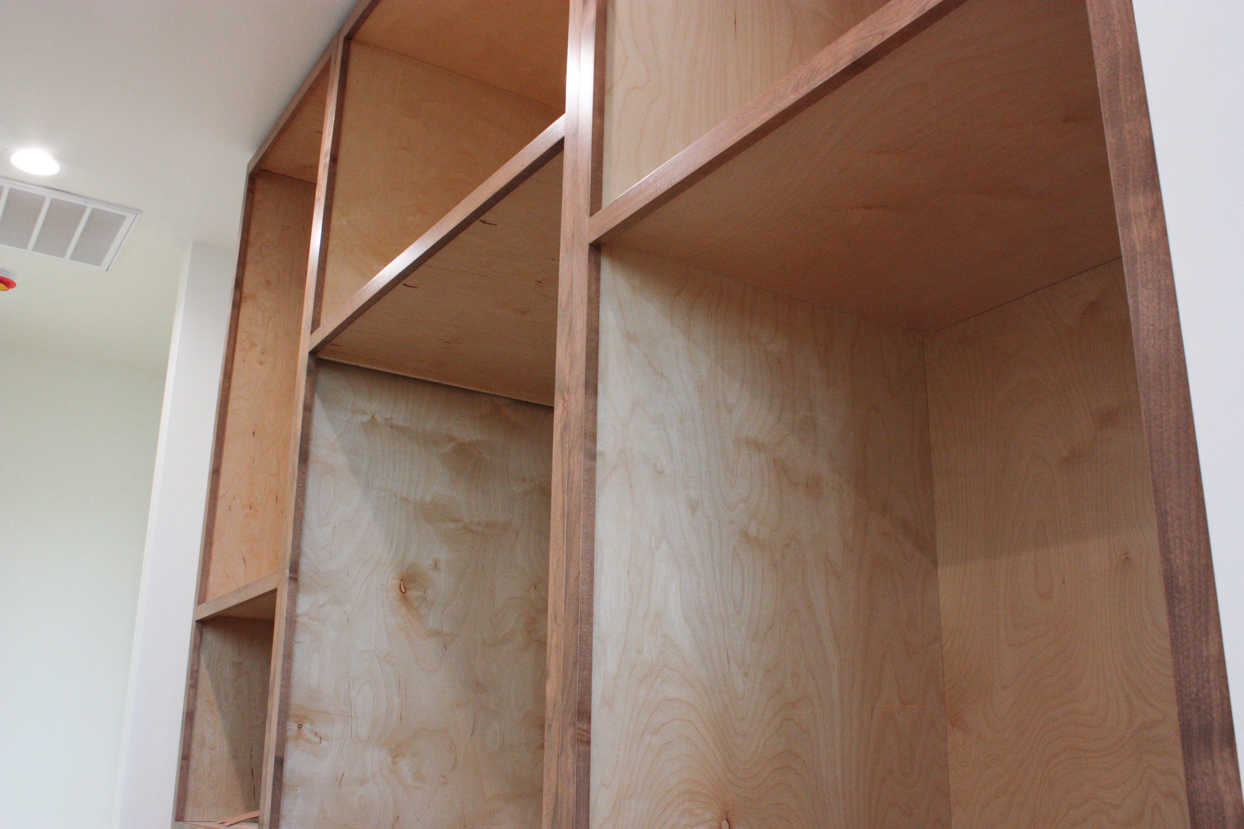 Unfinished wooden shelving unit in a room with white walls and ceiling, with a ceiling vent and a recessed light.