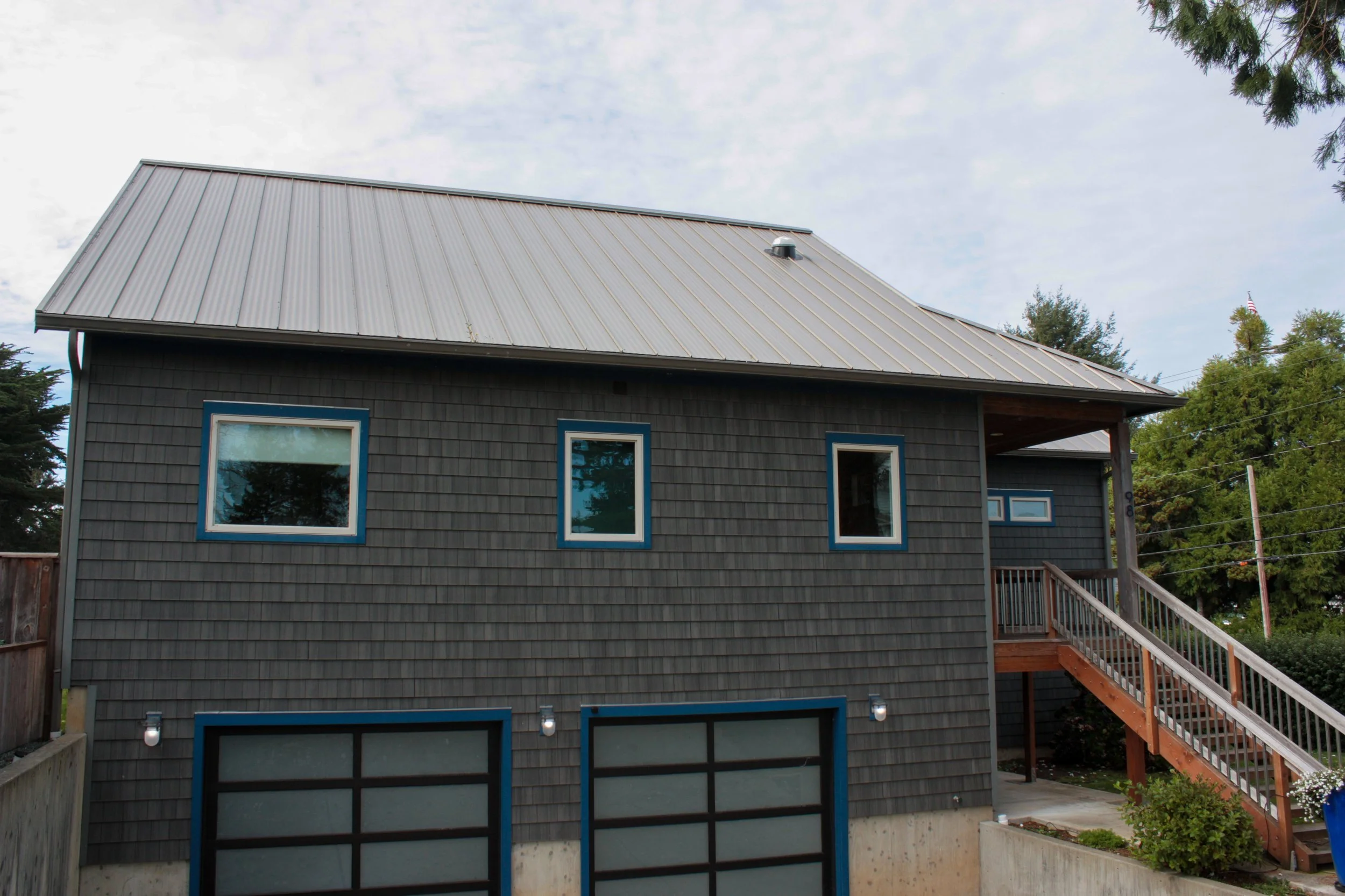 A two-story house with dark gray shingle siding, blue-trimmed windows, a metal roof, and an exterior staircase.