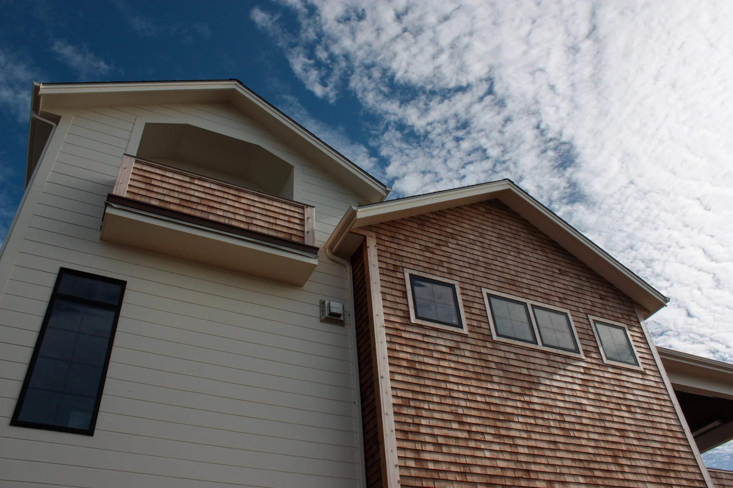 A modern two-story house with siding and wooden shingles, with several windows and a small balcony under a partly cloudy sky.