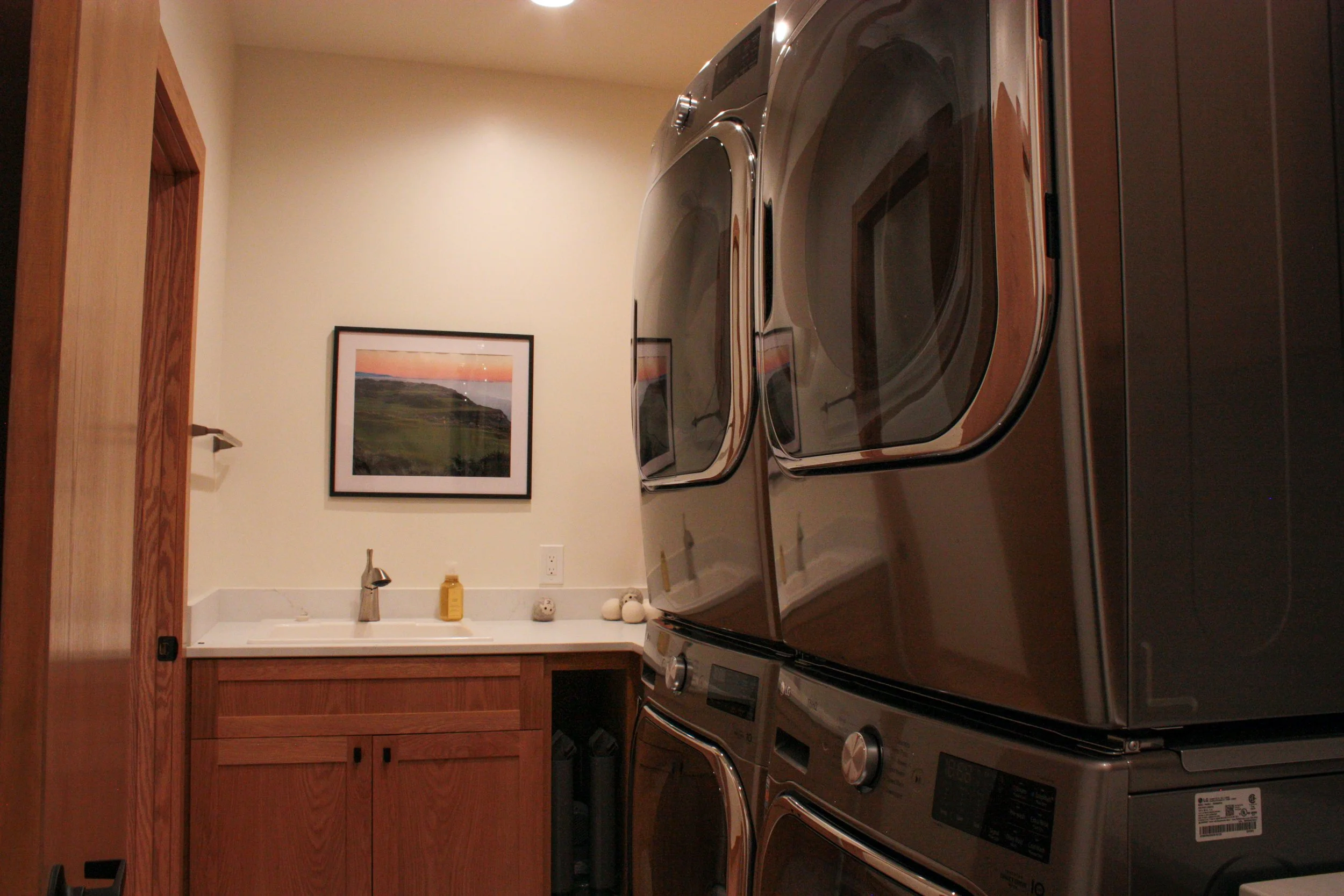 A laundry room with wooden cabinets, a white counter, a framed landscape photograph on the wall, and a stack of metallic washing machines and dryers.