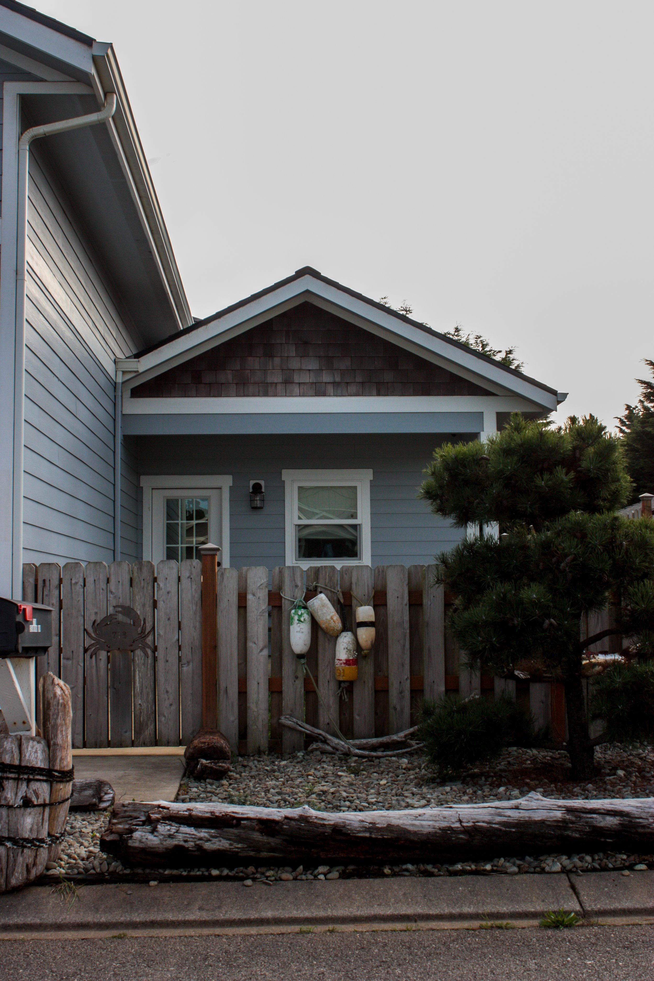 Front of a blue house with a small porch, surrounded by a wooden fence with nautical decor, including buoy floats and a crab decoration, and a small tree in the yard.