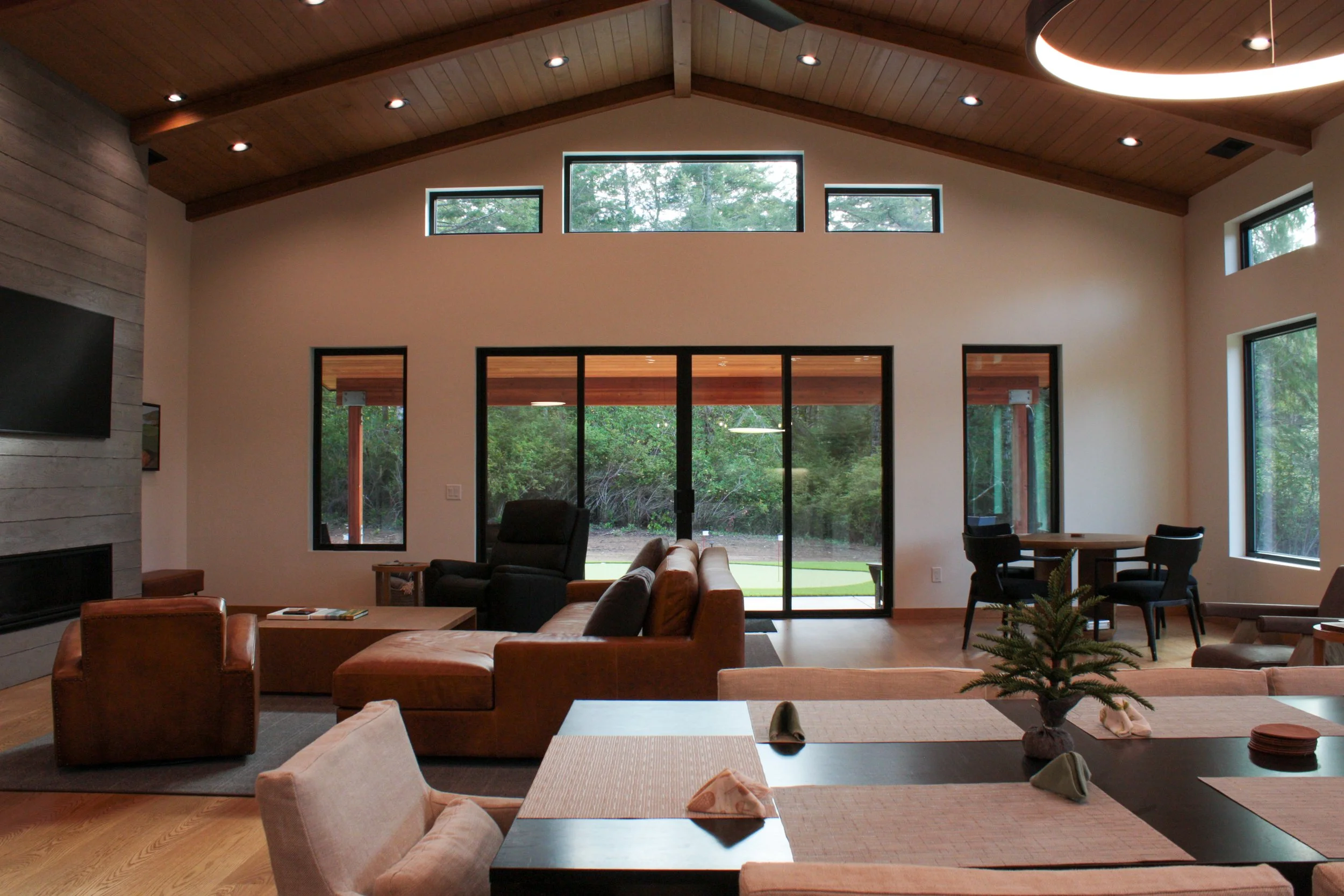 Modern living room with high vaulted ceiling, large windows, sliding glass door, and wooden furnishings, opening to a backyard with trees.