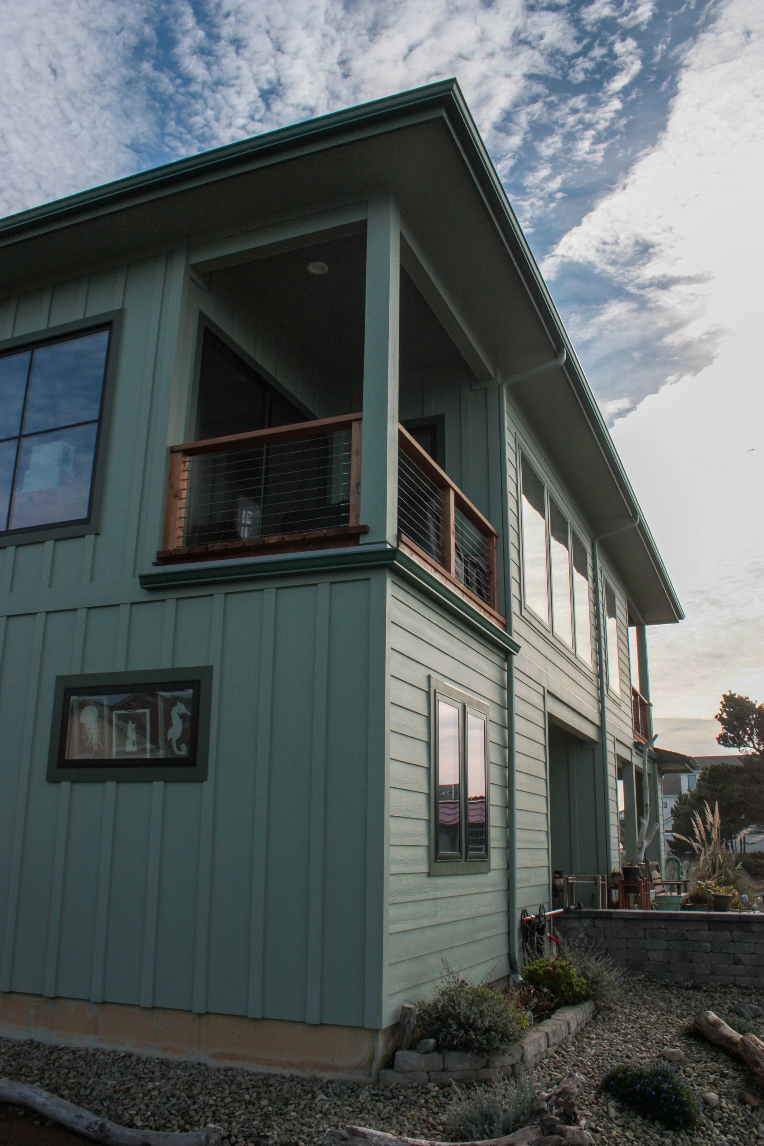 A two-story modern house painted light blue with large windows, a small balcony with wooden railing, and a landscaped yard with plants and rocks.