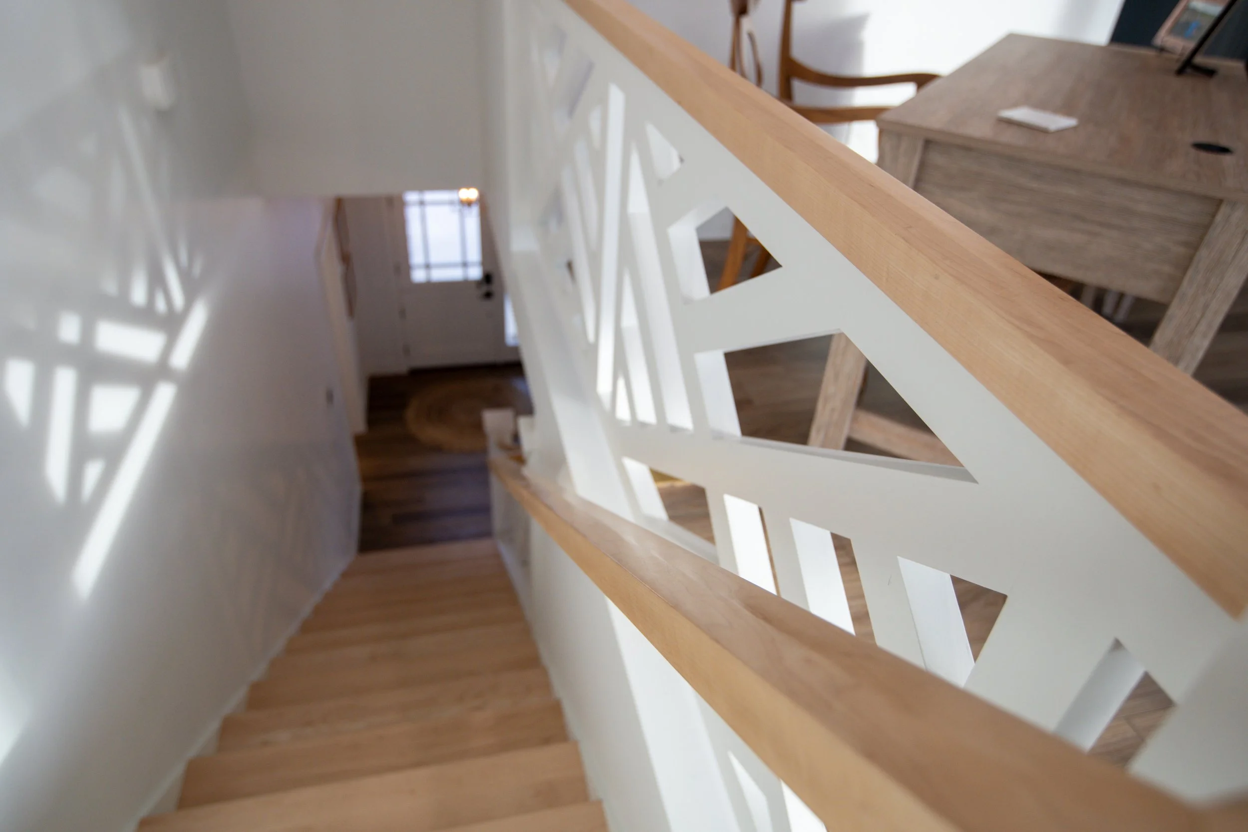View of a wooden staircase from the top, showing the railing with geometric cutouts, leading down to a hallway with hardwood flooring and a white door at the end.