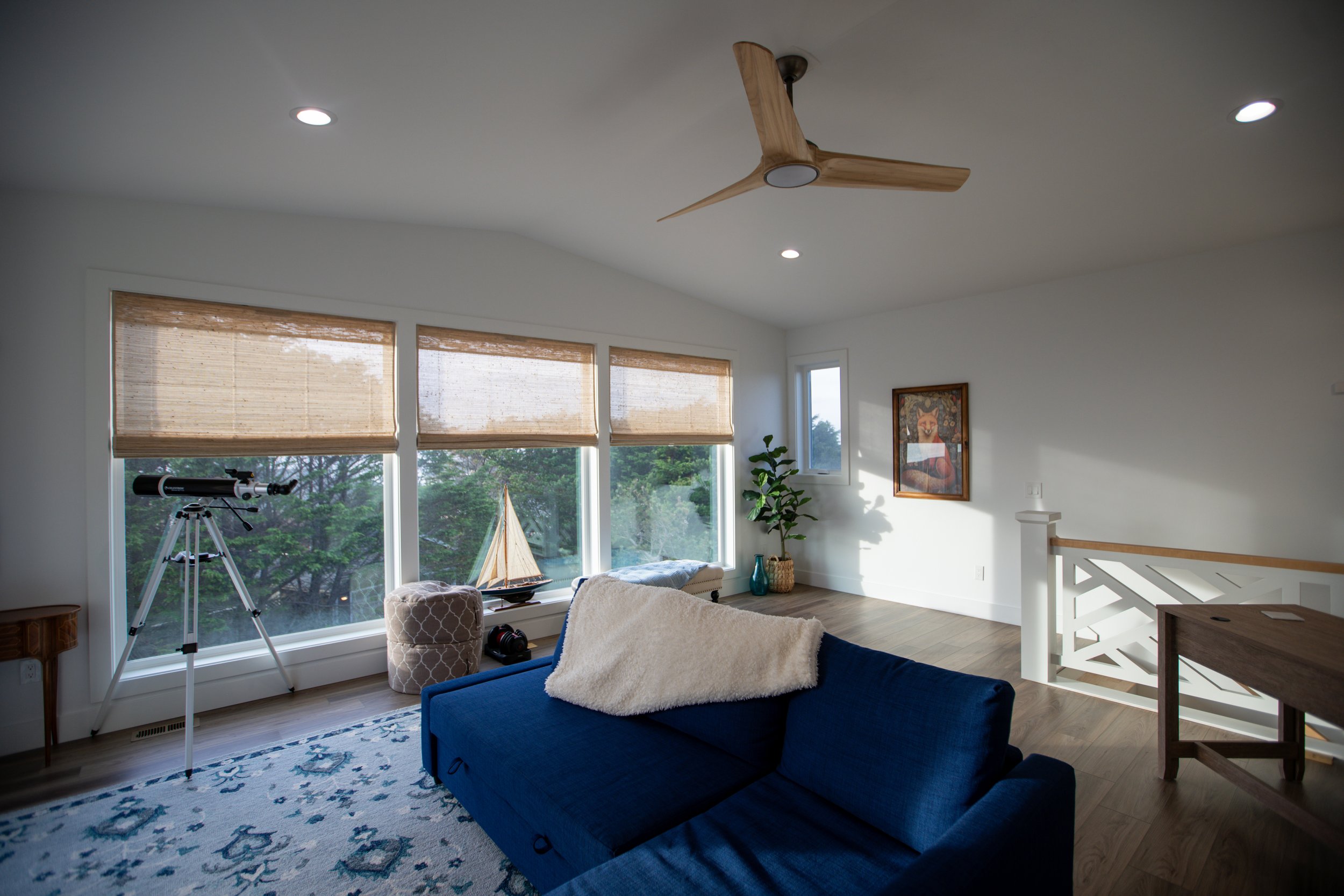 Living room with large front windows, blue sofa, telescope near window, sailboat model, potted plant, framed artwork, ceiling fan, and wood flooring.
