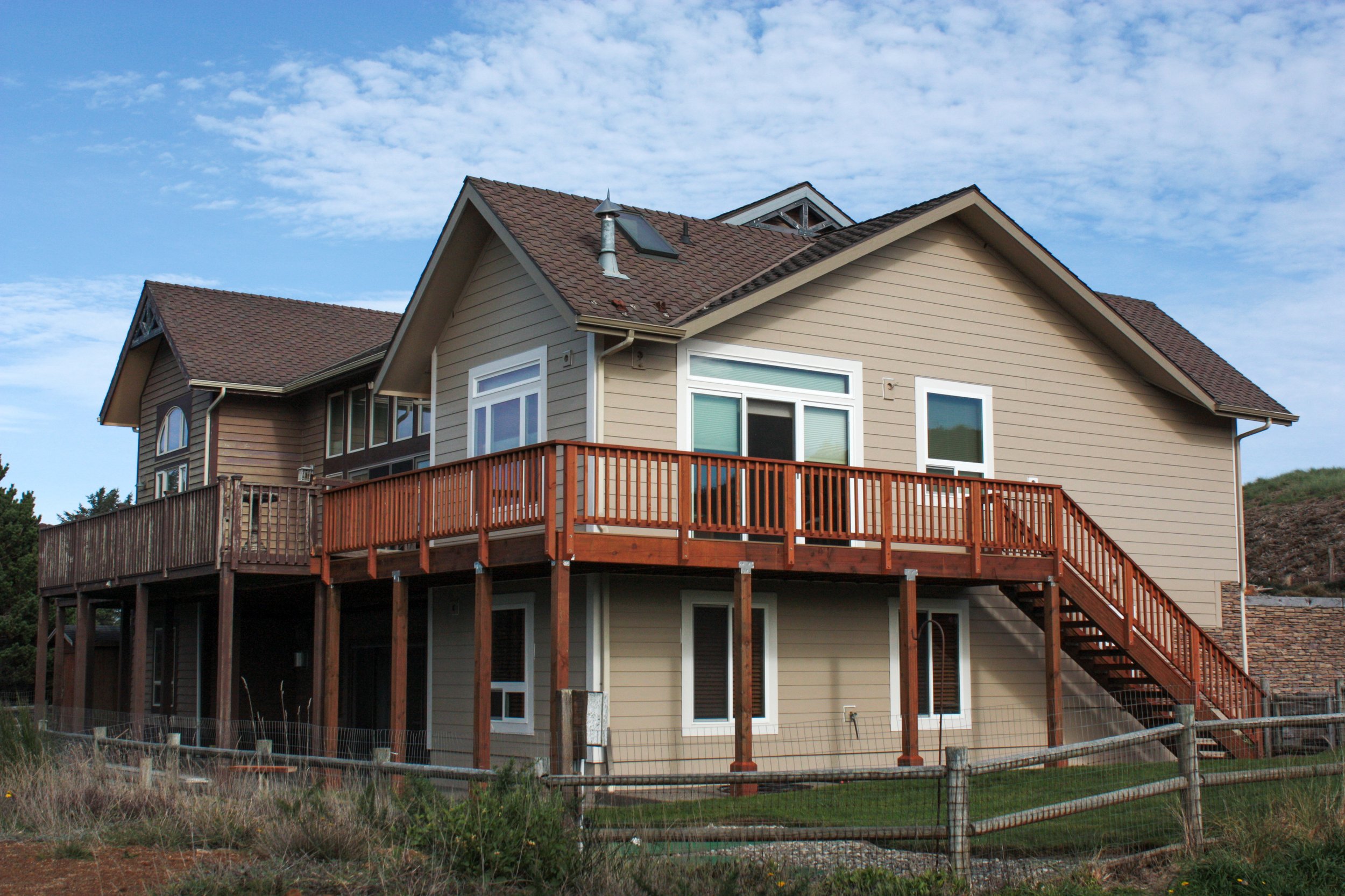 A multi-story house with beige siding, brown roofing, and a large wooden deck on the second level with stairs leading down.