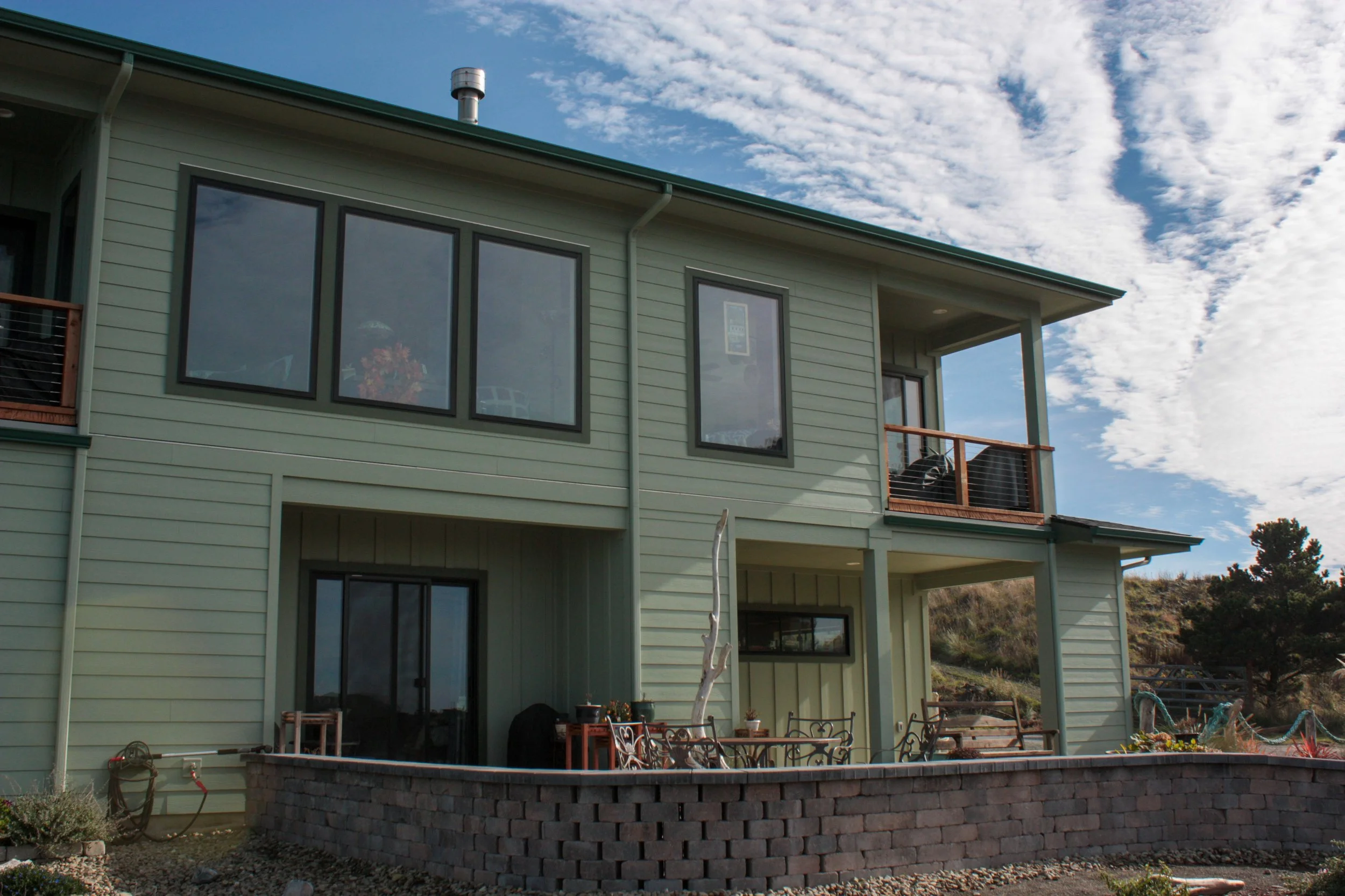 A two-story green house with large windows and a small outdoor patio with chairs and tables, set against a partly cloudy sky and a hillside landscape.