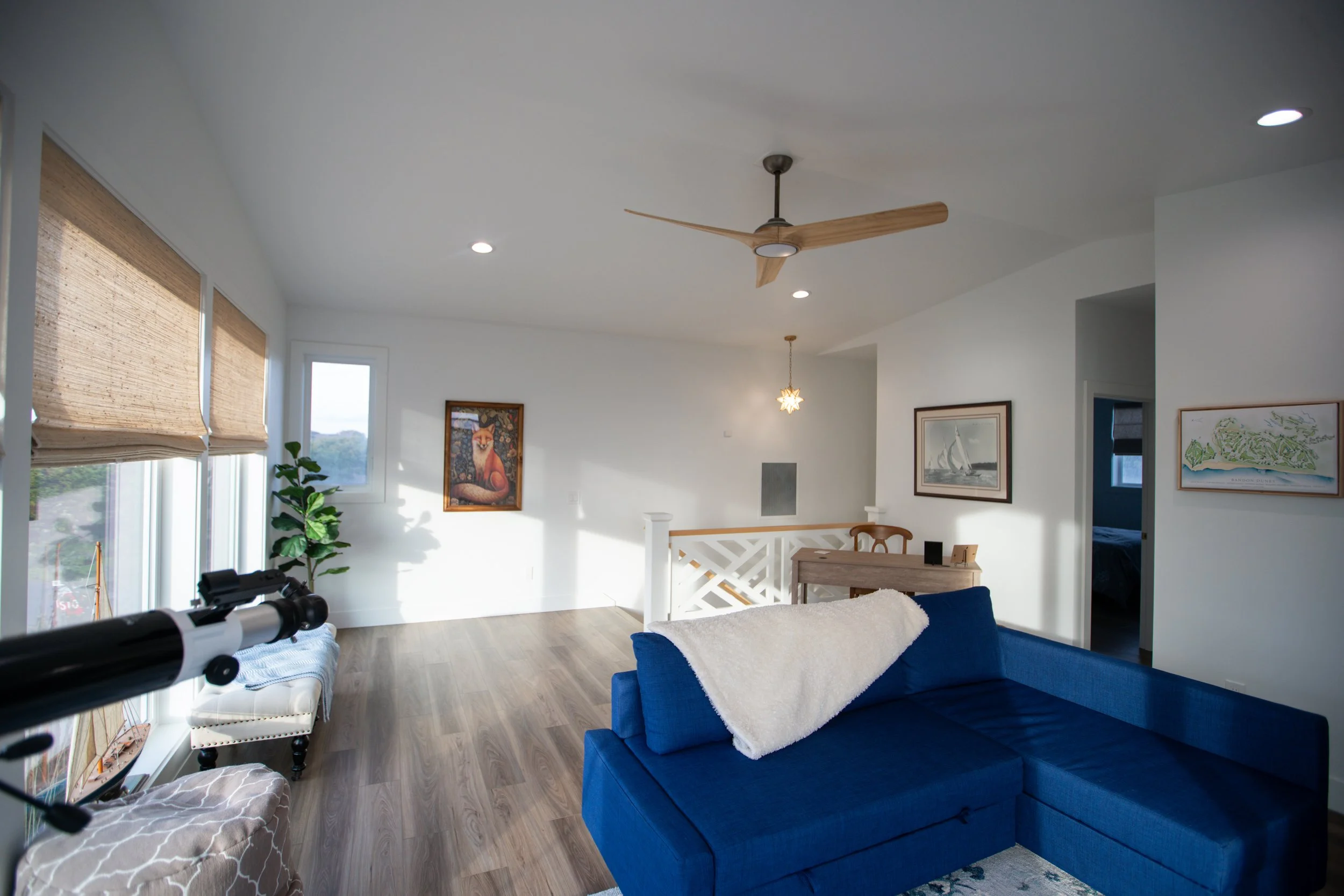 Living room with white walls, large windows with bamboo blinds, a blue sectional sofa with a white blanket, wooden floors, artwork on the walls, a telescope near the window, and a small desk with a wooden chair.