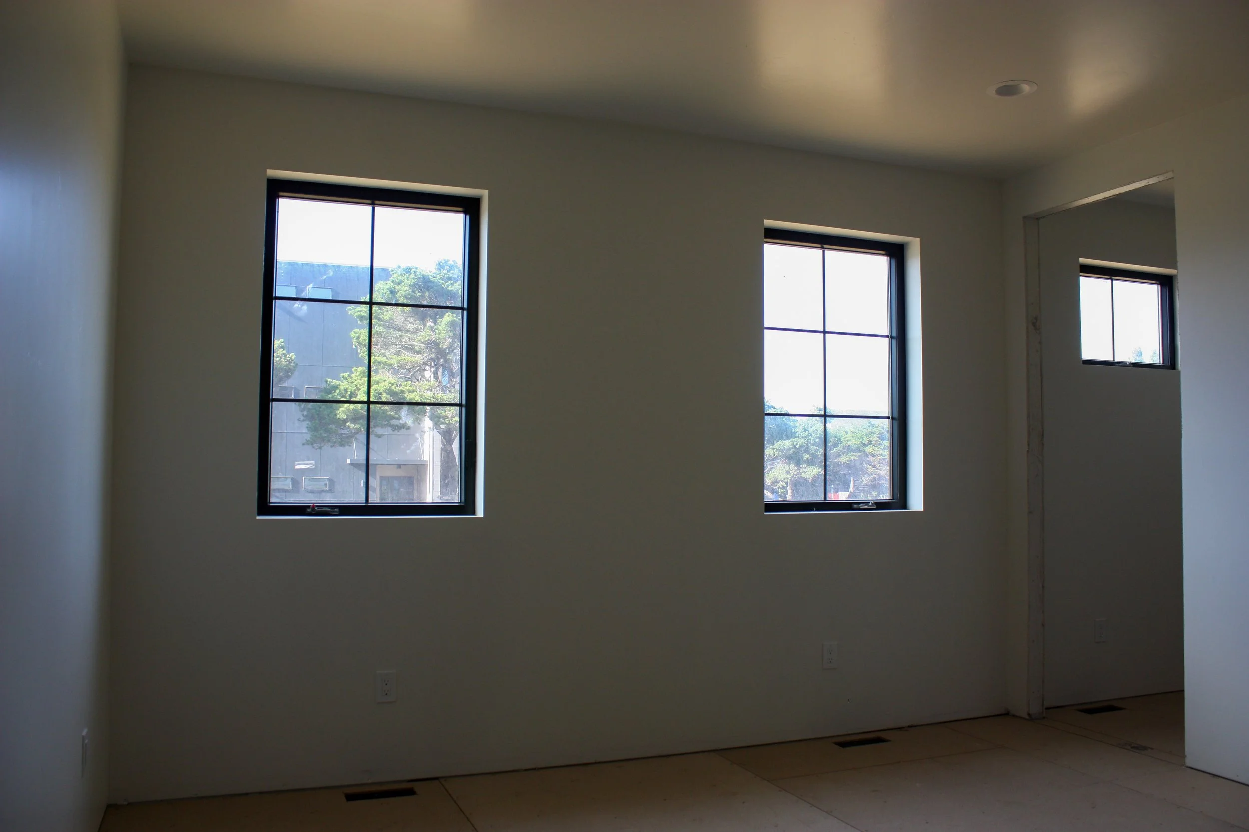 Empty room with three windows showing trees outside, white walls, and a beige floor.