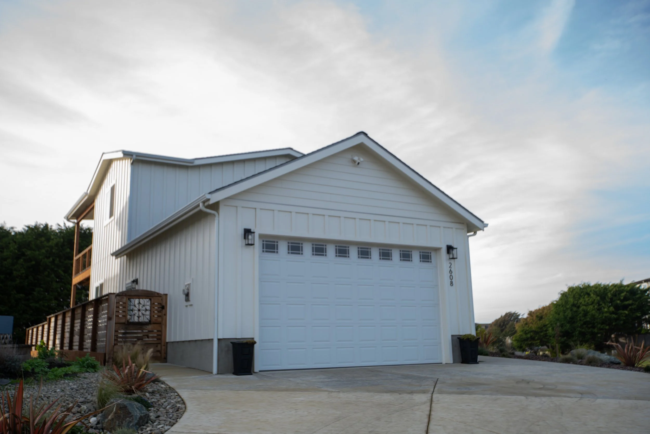 A white two-story house with a garage, a wooden deck, and a landscaped front yard with plants and rocks.