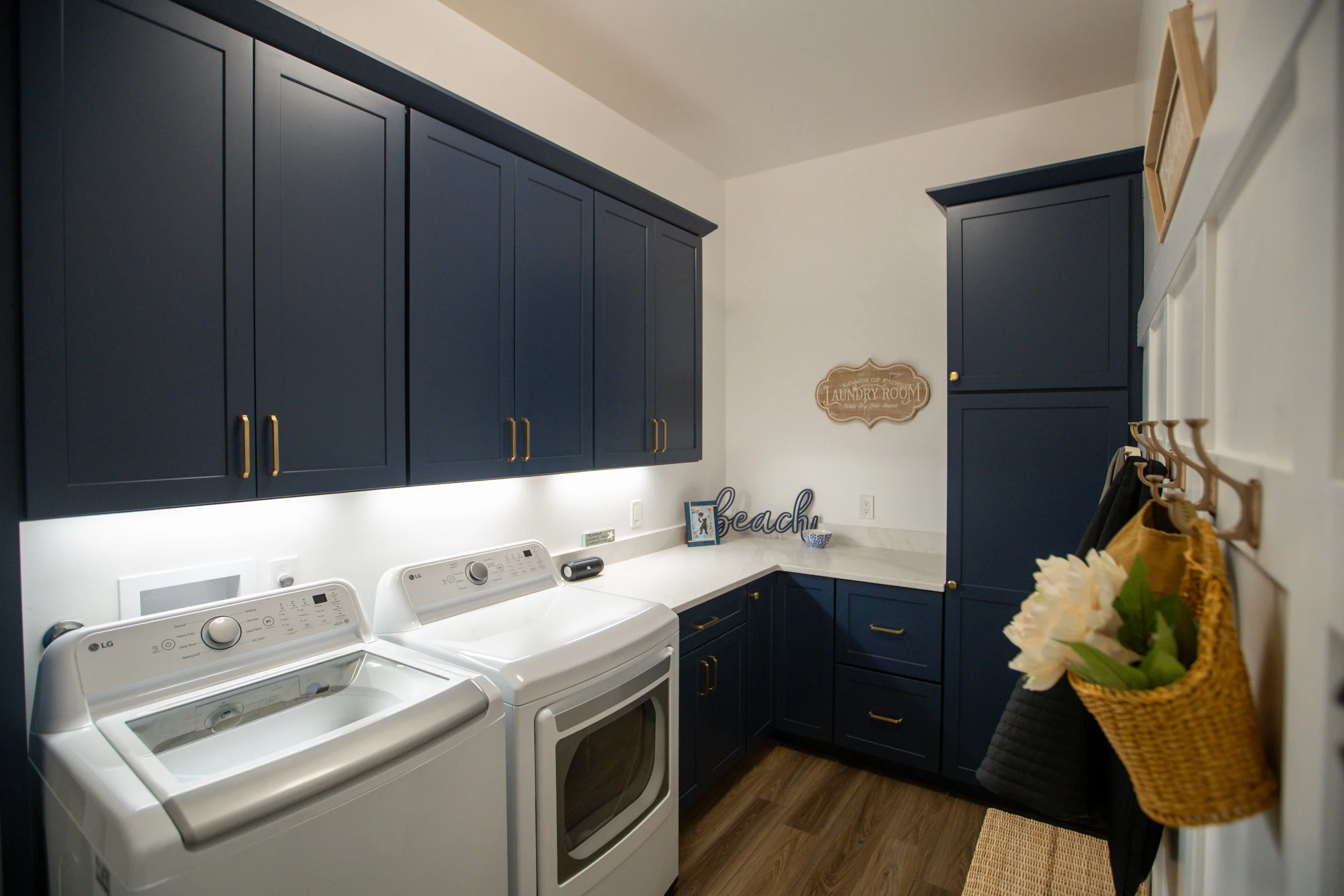 Laundry room with blue cabinets, white washer and dryer, wall sign reading 'Laundry Room,' and a wall hook with a basket of flowers and hanging towels.