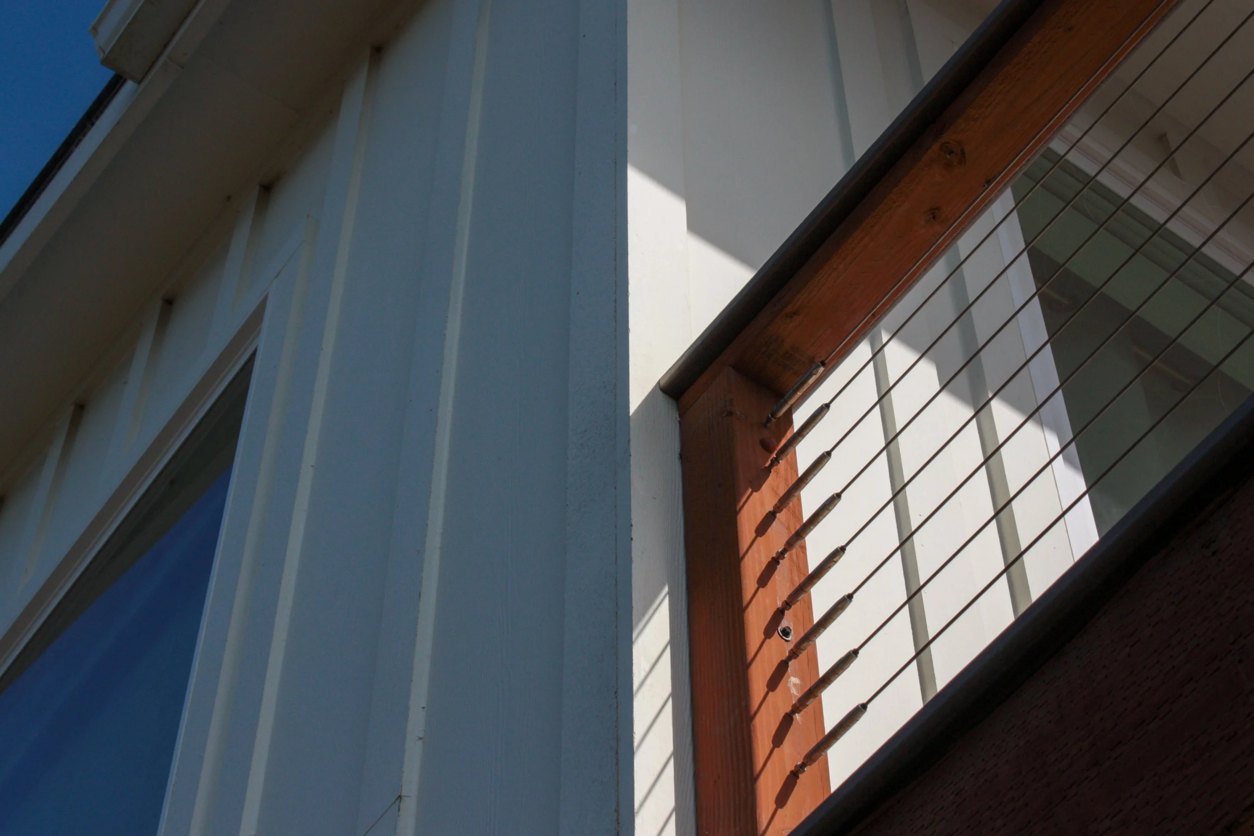 Close-up of a balcony with wooden railings and wire cables, part of a modern building with white exterior walls and large glass windows.
