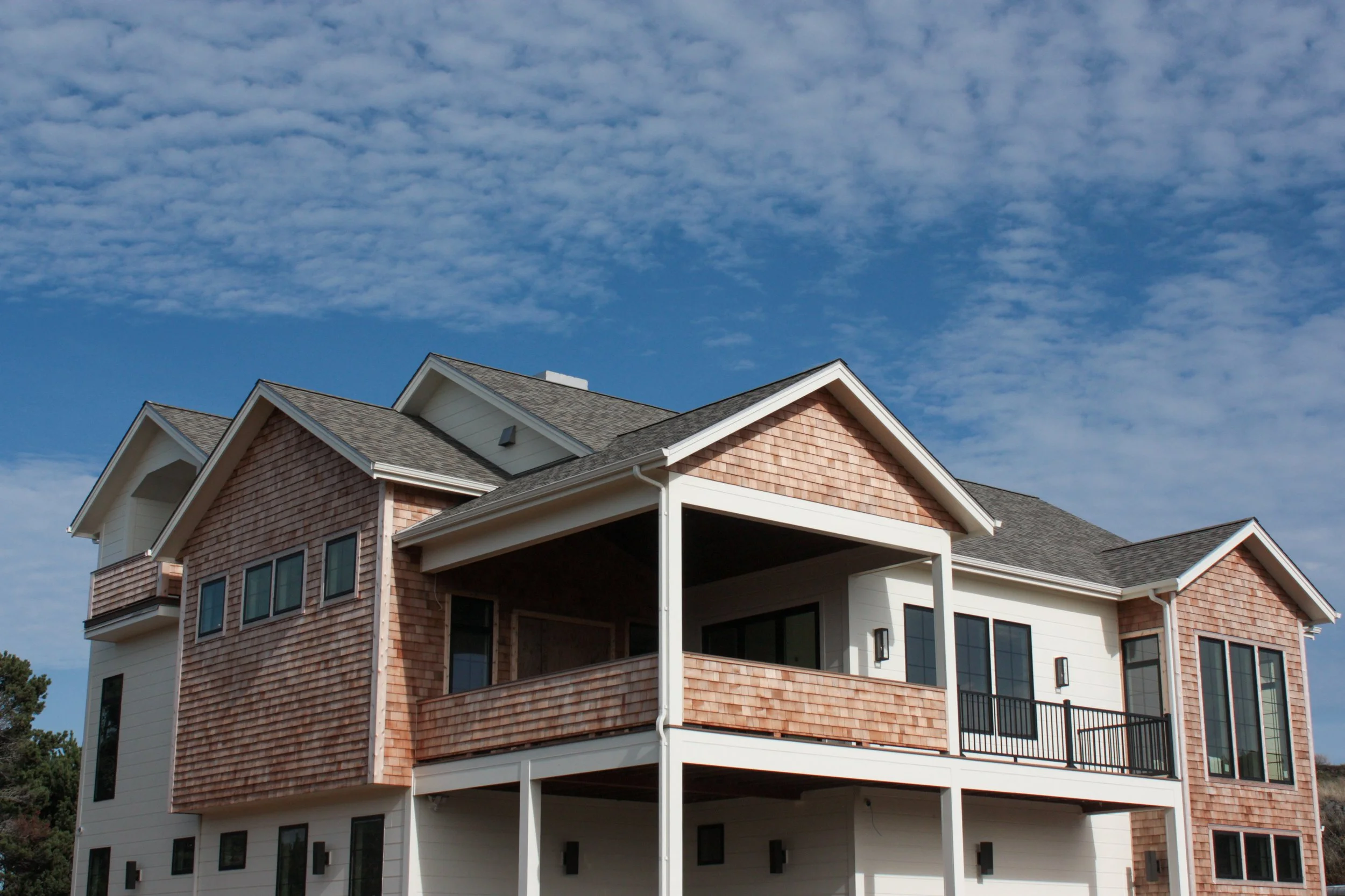 A modern, multi-story house with a combination of shingle and siding exterior, large windows, a balcony with black railing, and a sloped roof, under a partly cloudy blue sky.