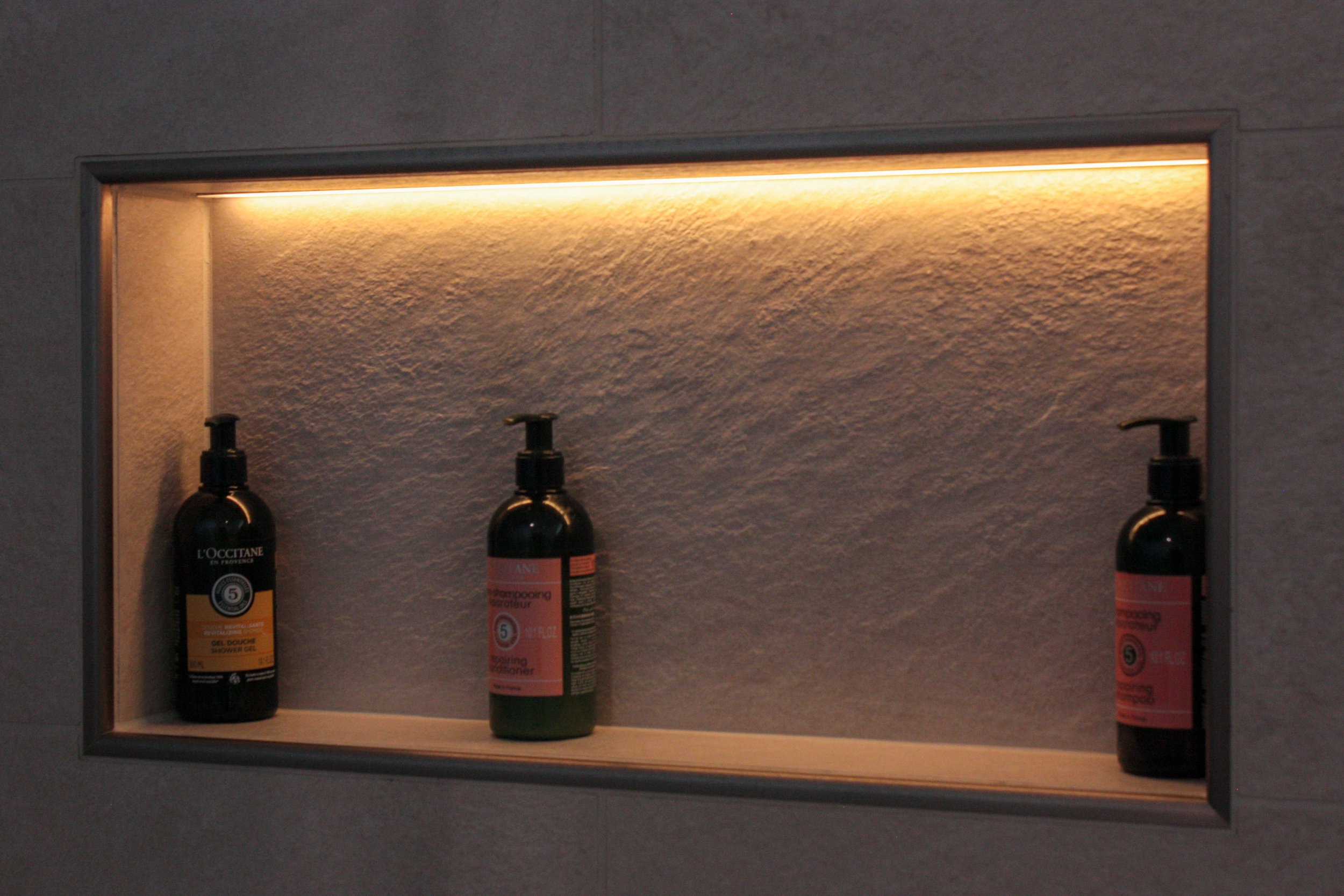 Bathroom wall niche with three bottles of L'Occitane shower and shampoo gels on a shelf, illuminated with warm lighting.