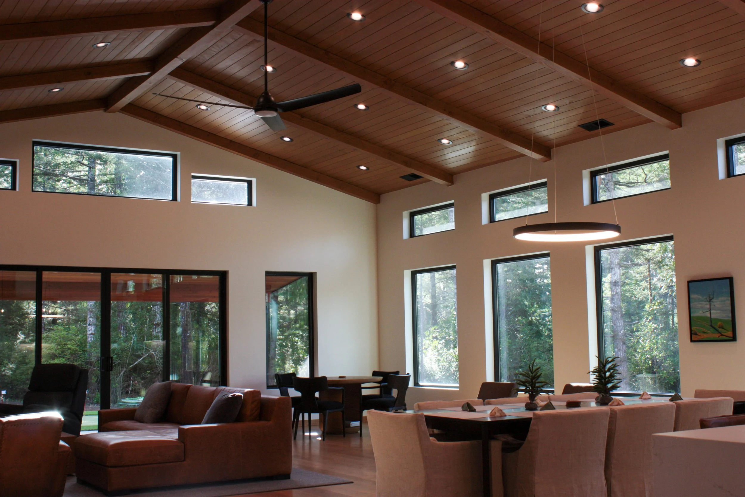 Interior view of a modern living and dining area with large windows, wooden ceiling with recessed lighting, ceiling fan, and contemporary furniture.