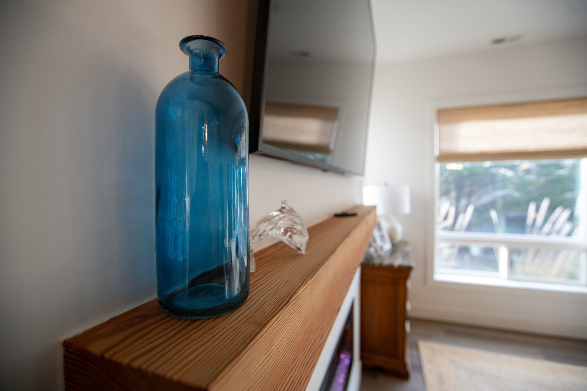 A blue glass bottle on a wooden mantelpiece in a living room with a large window and a TV mounted on the wall.