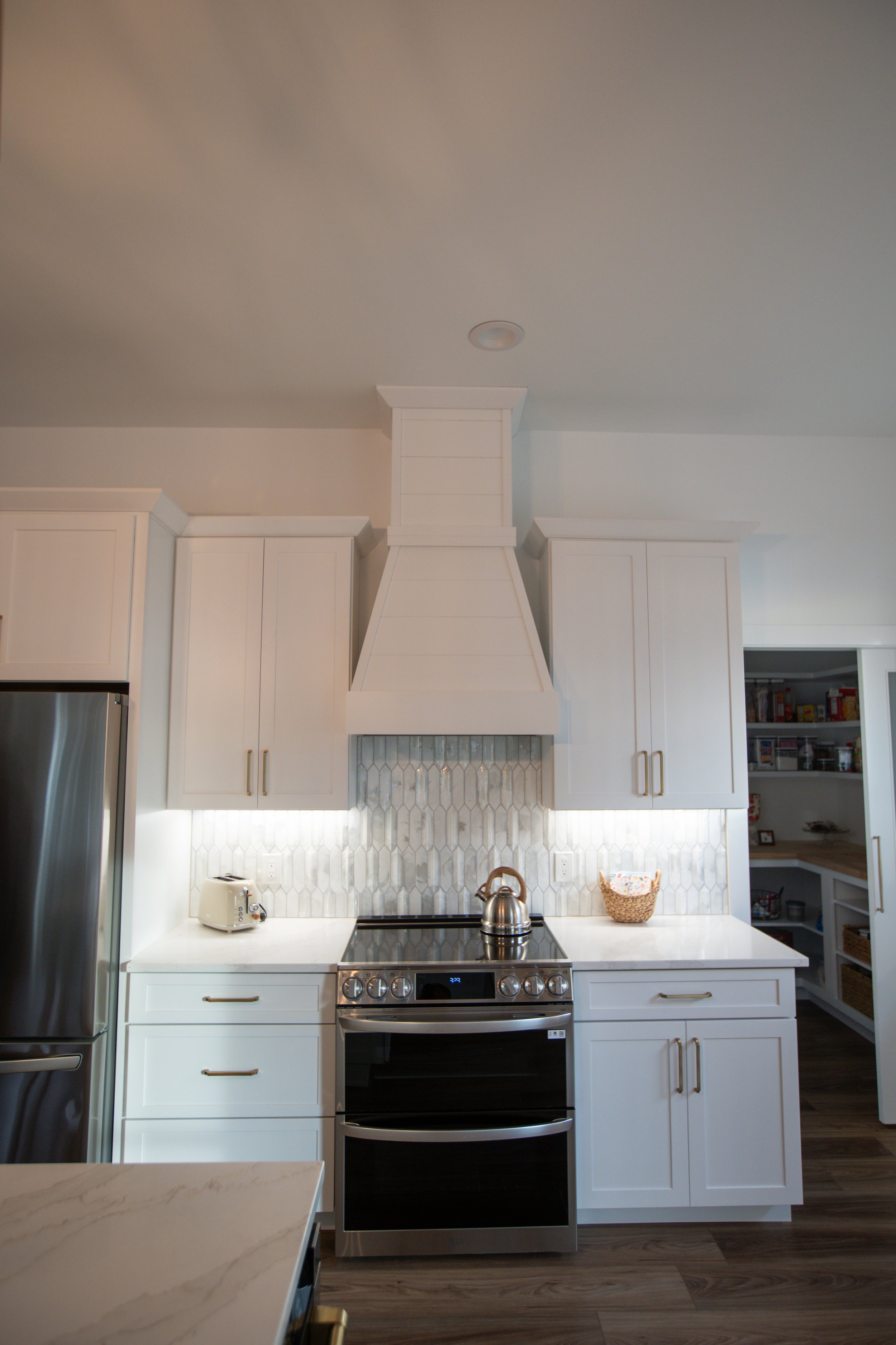 Modern kitchen with white cabinets, stainless steel refrigerator, and a range with a teapot on top. There are light-colored countertops, a marble backsplash, and a pantry with open shelves in the background.